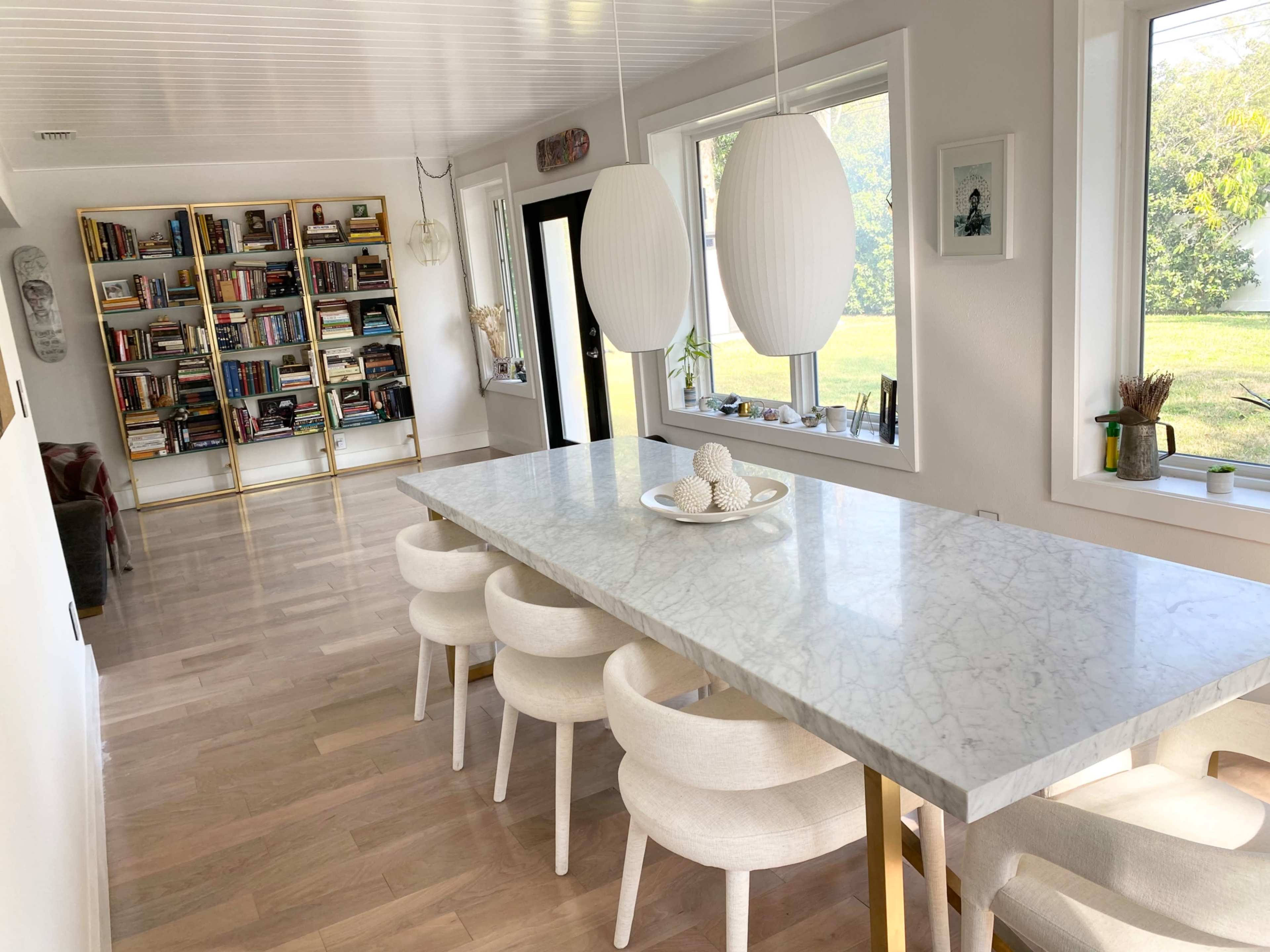 A modern dining area features a marble table with round white chairs, surrounded by large windows and a bookshelf filled with books.