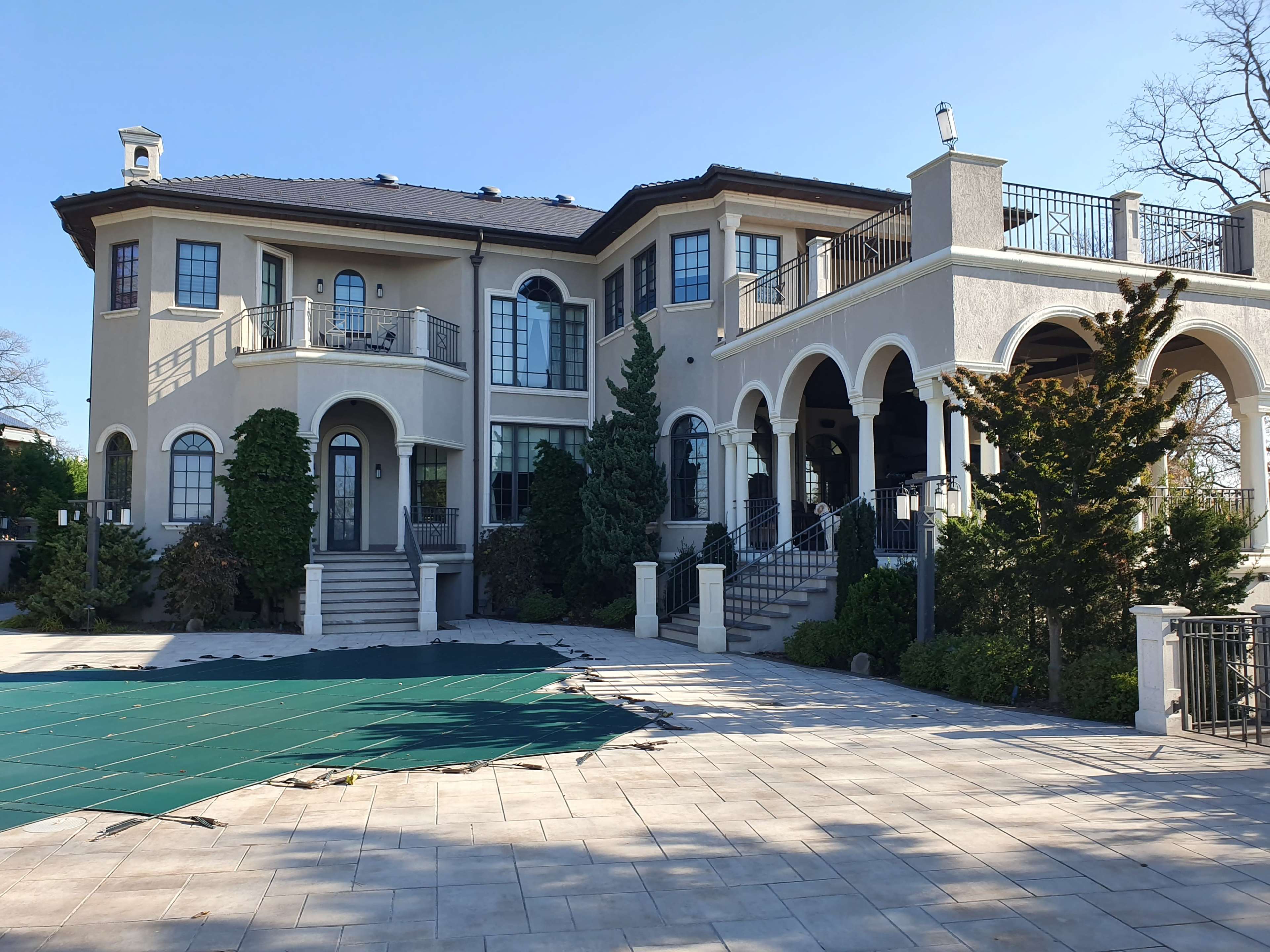A large, two-story house with a mix of arched and rectangular windows, a balcony, and a stone walkway, set against a clear sky.