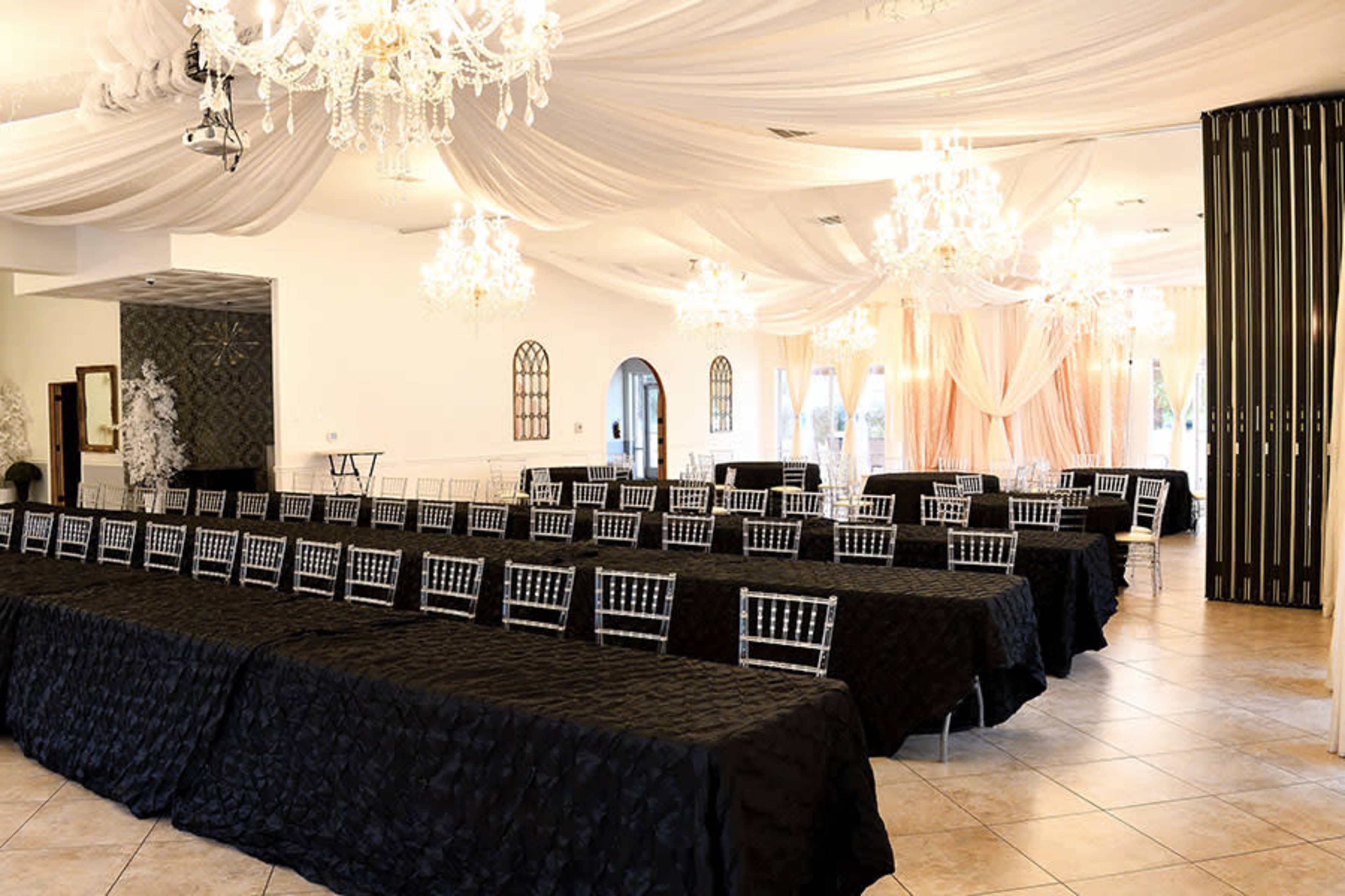 A banquet hall arranged with rows of tables covered in black tablecloths and adorned with silver Chiavari chairs under draped fabric and chandeliers.