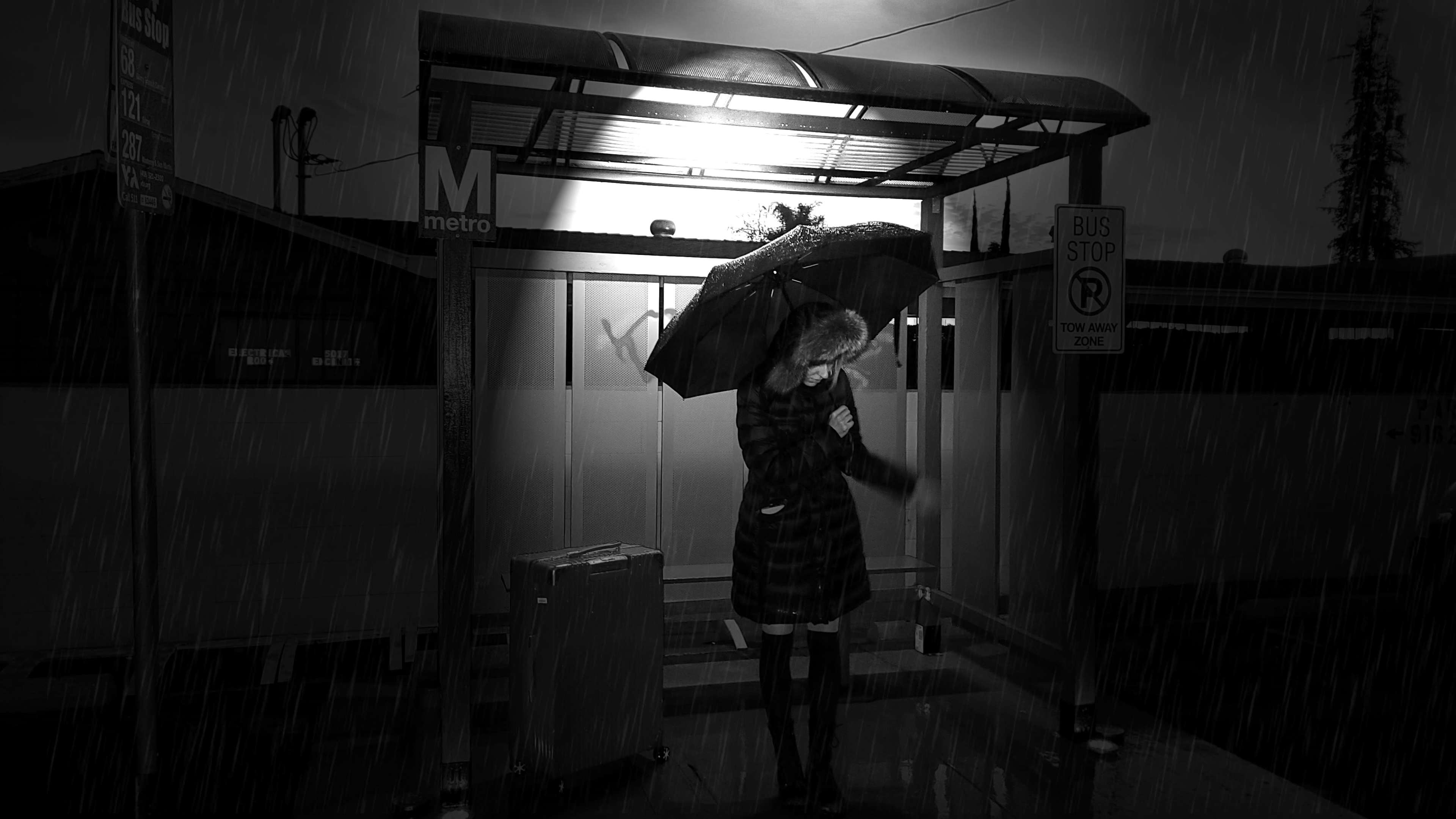 A person holding an umbrella stands at a bus stop in the rain, next to a suitcase, under a lit shelter.