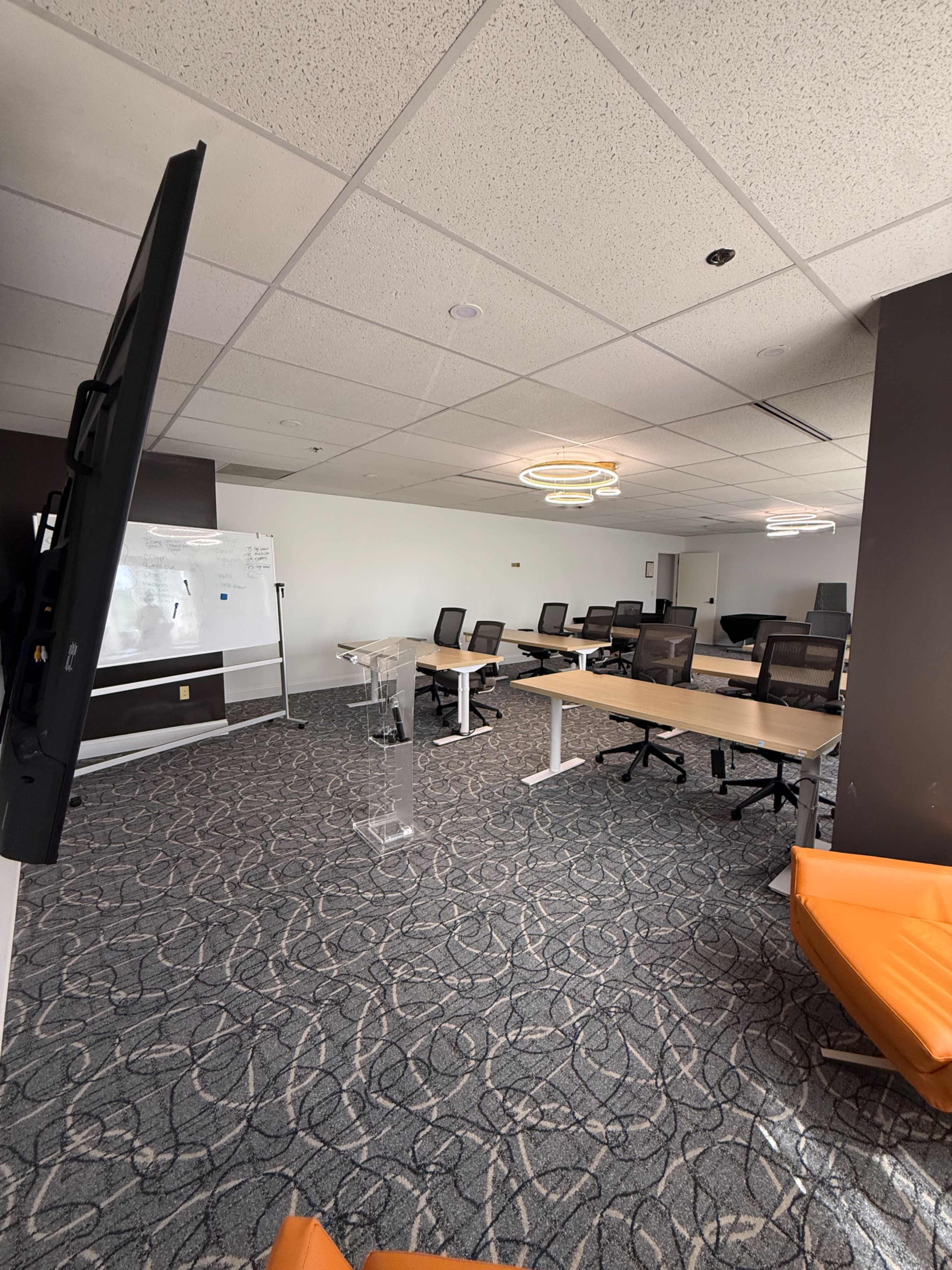 The image shows a modern, open office space featuring several desks, ergonomic chairs, a large screen, and a clear floor layout with a patterned carpet.