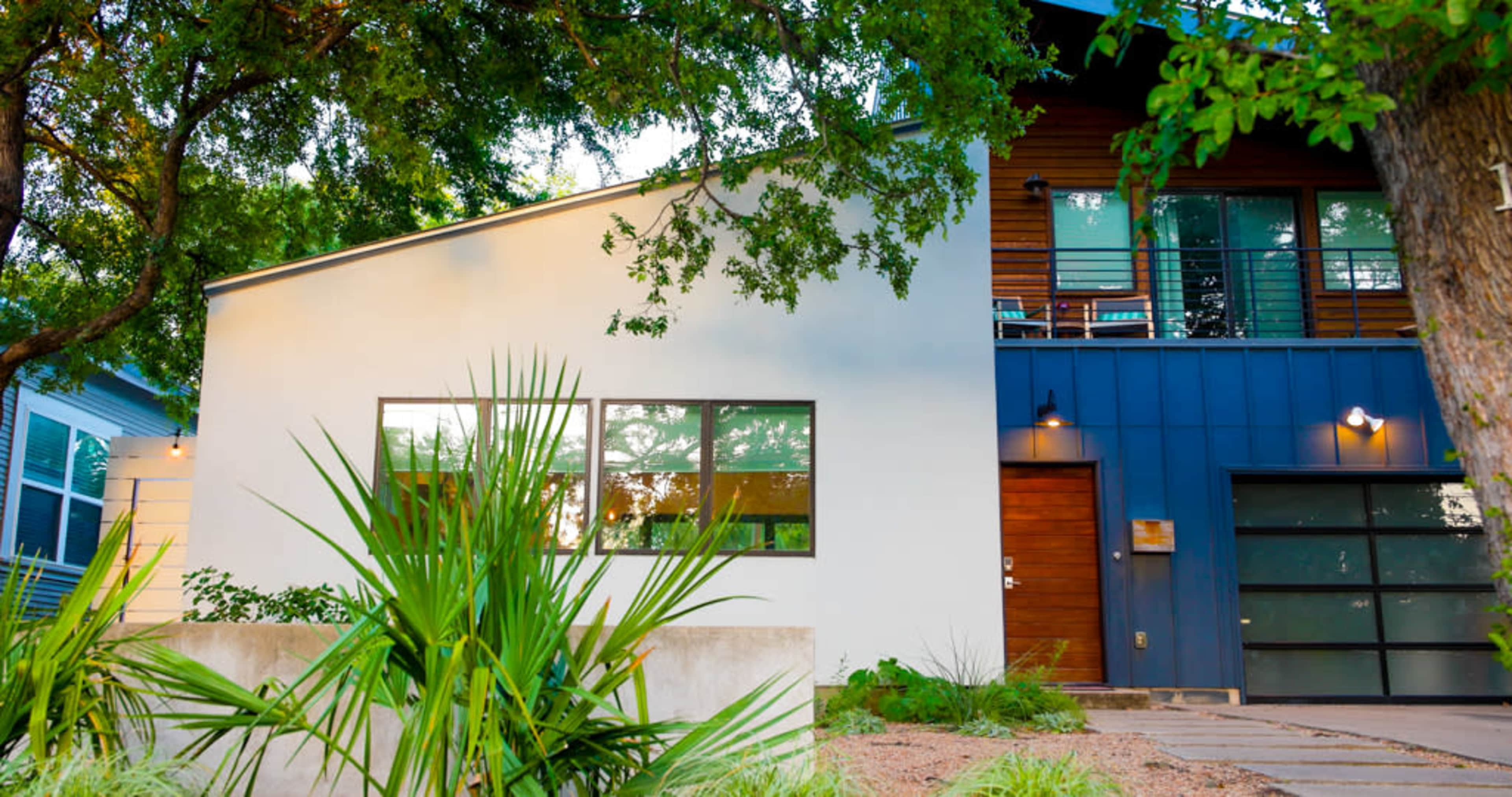 The image shows a modern two-story house with a mix of white and blue siding, surrounded by greenery and featuring large windows.