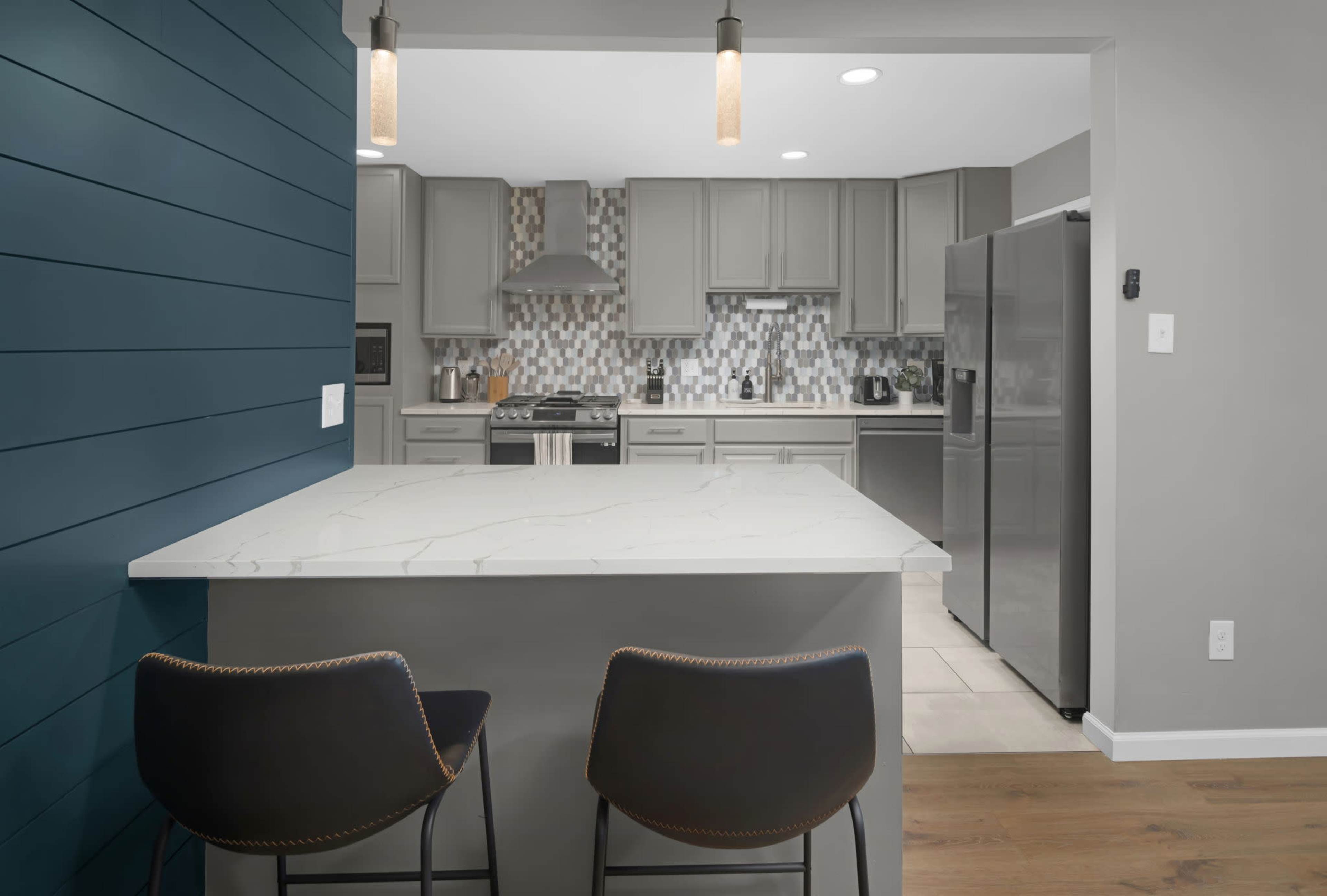 The image shows a modern kitchen with gray cabinets, a mosaic tile backsplash, and bar stools at a marble countertop.