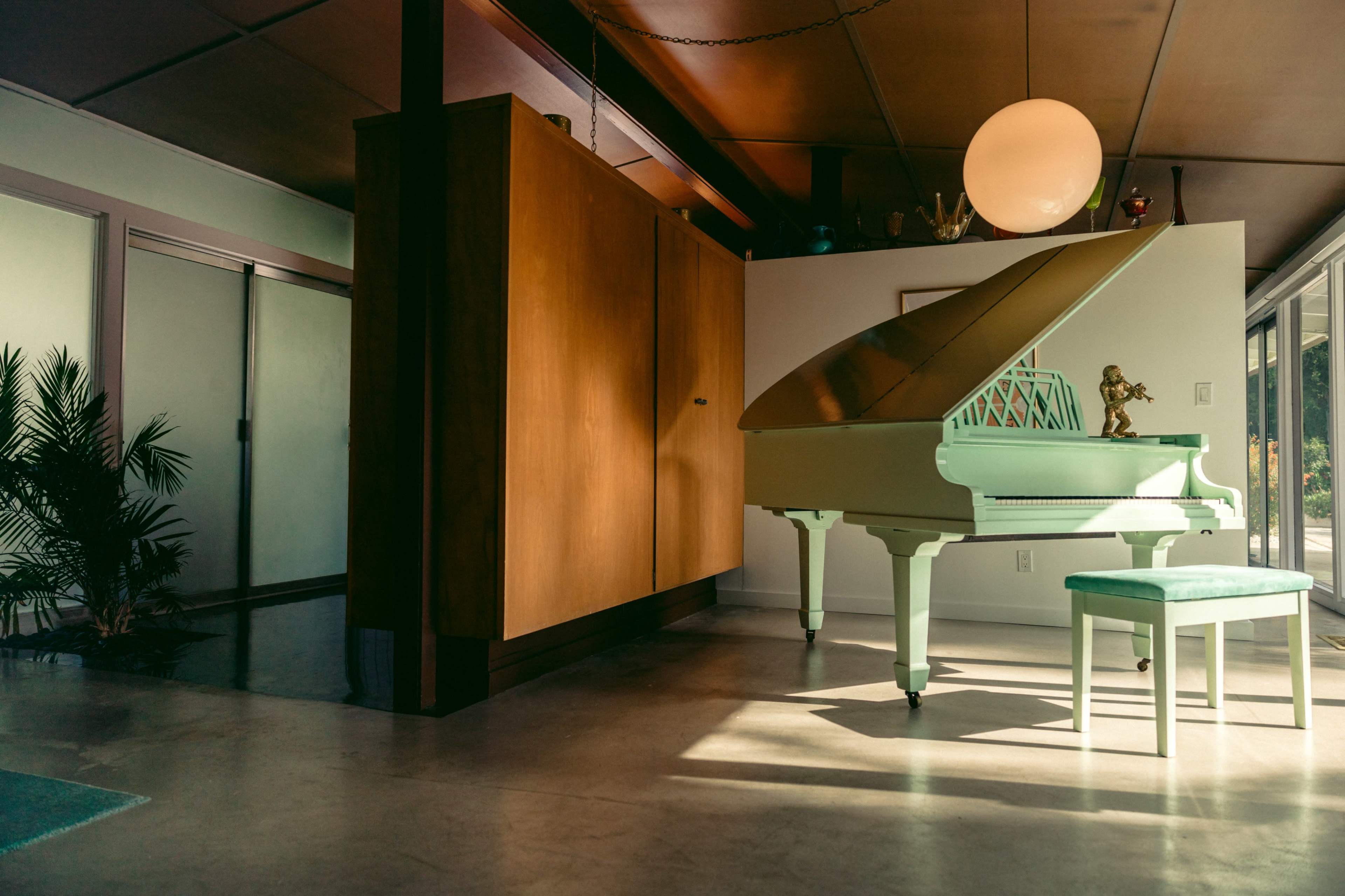A grand piano in a light green finish is positioned next to a wooden wall unit in a modern interior space with large windows.