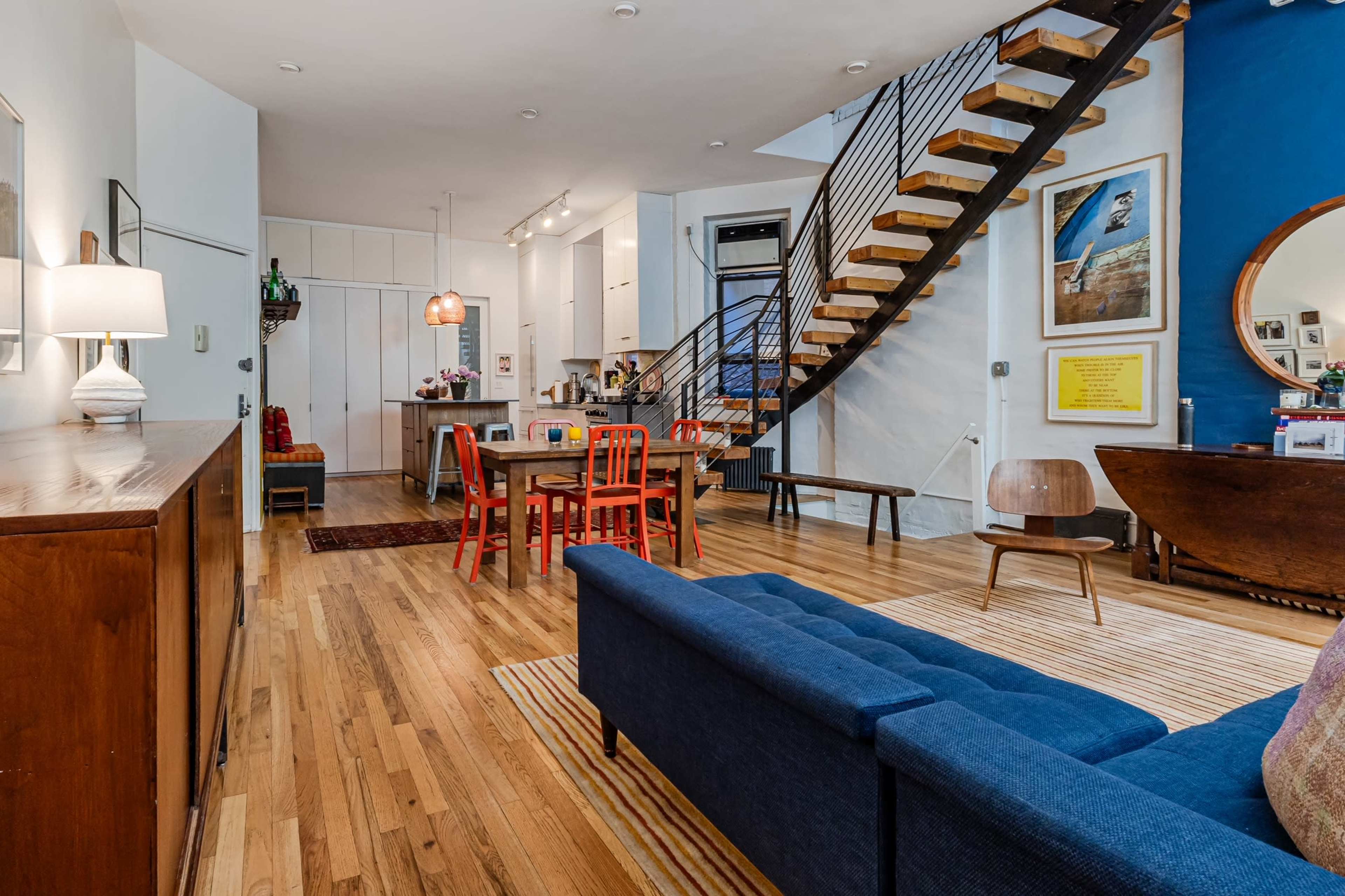 A modern living space with a blue sofa, a wooden dining table surrounded by red chairs, a staircase leading to an upper level, and various artworks on the walls.