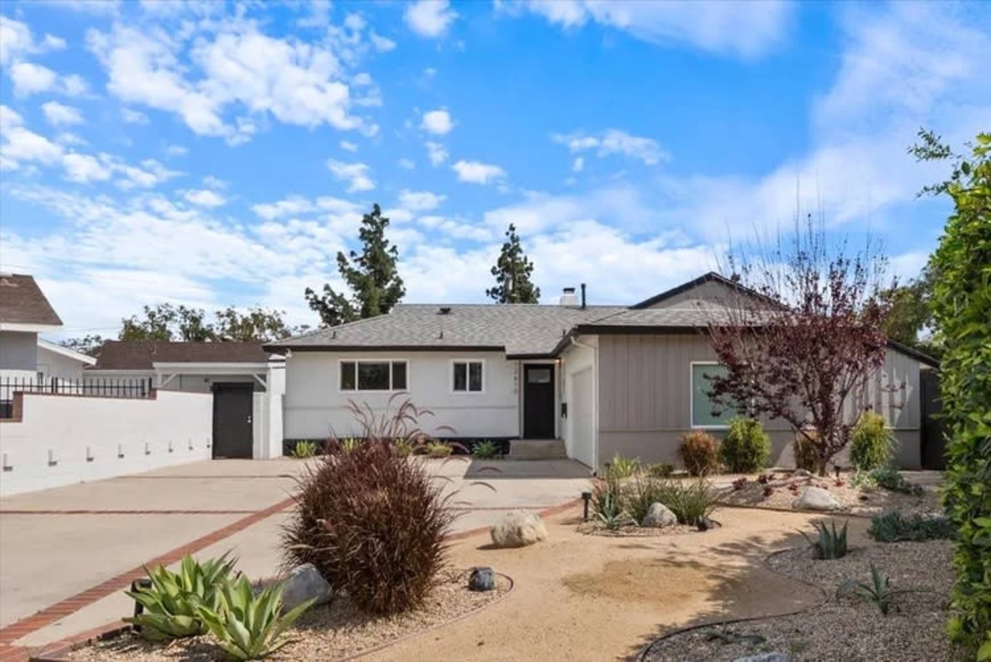 A single-story house with a landscaped front yard, gravel driveway, and various plants is set against a blue sky with scattered clouds.