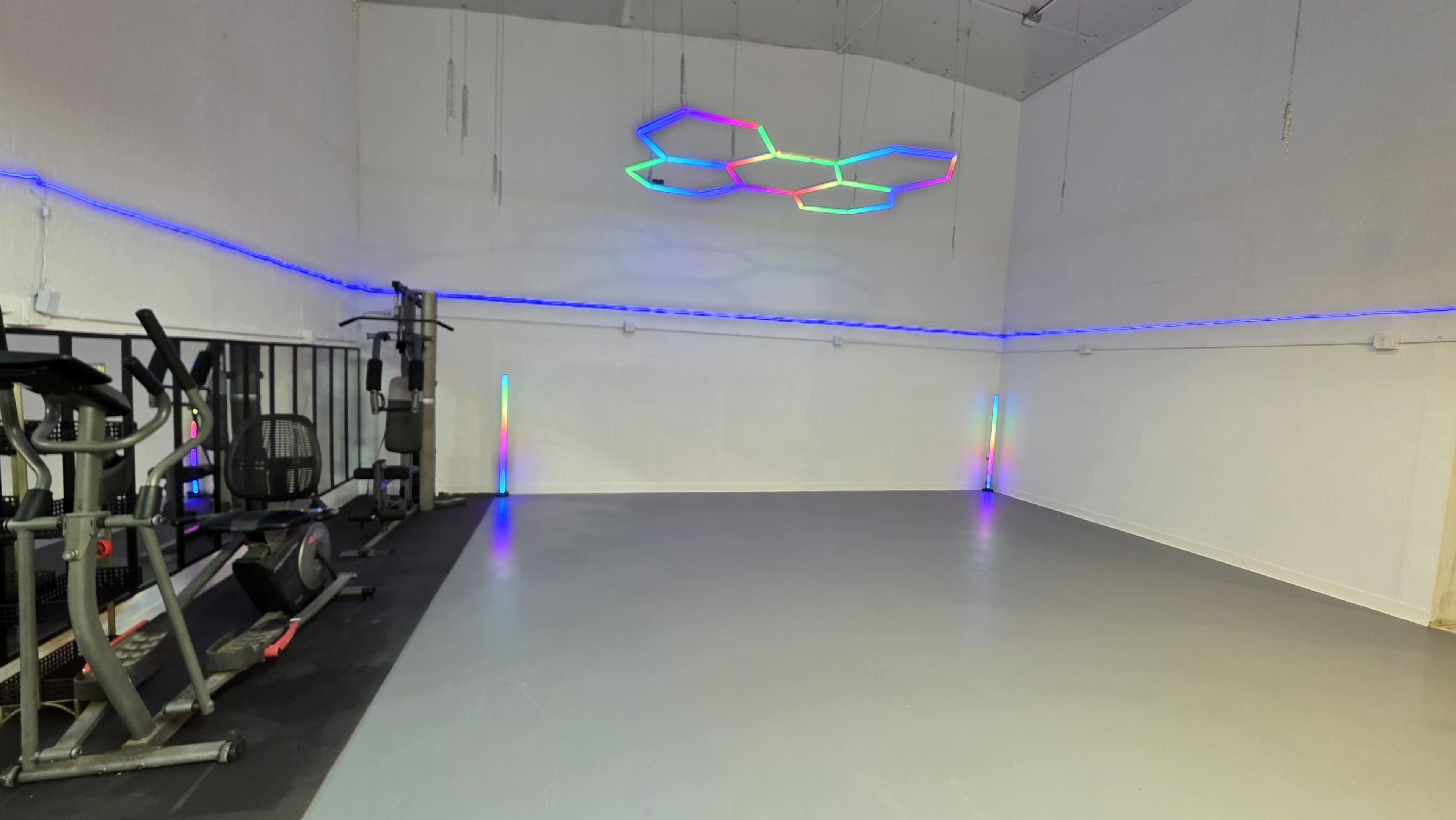 The image shows an empty gym space featuring exercise equipment on one side and colorful LED lighting along the walls and ceiling.
