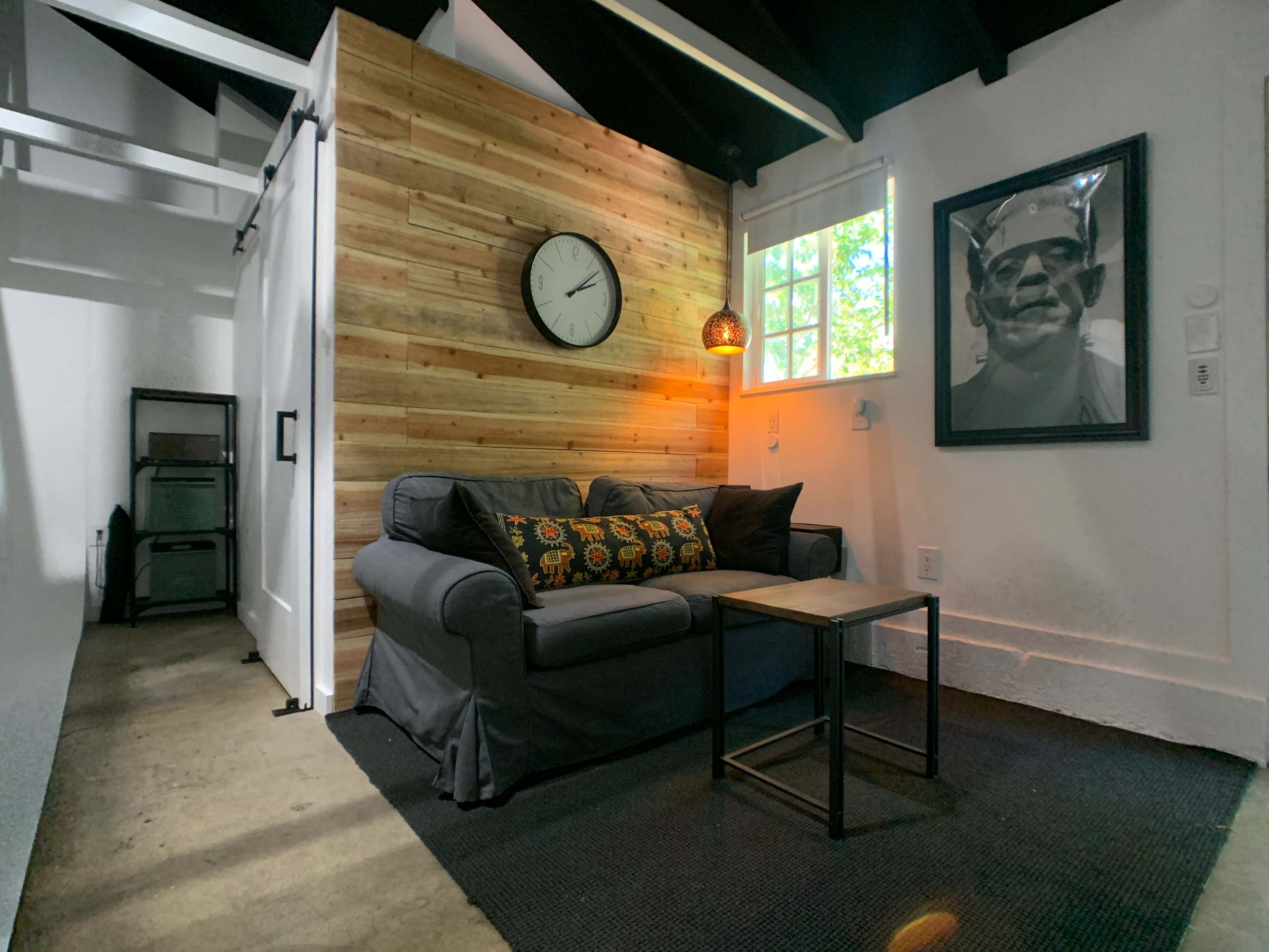 The scene features a cozy living area with a gray sofa adorned with colorful pillows, a small table beside it, a wooden accent wall with a clock, and a large framed portrait hanging nearby.