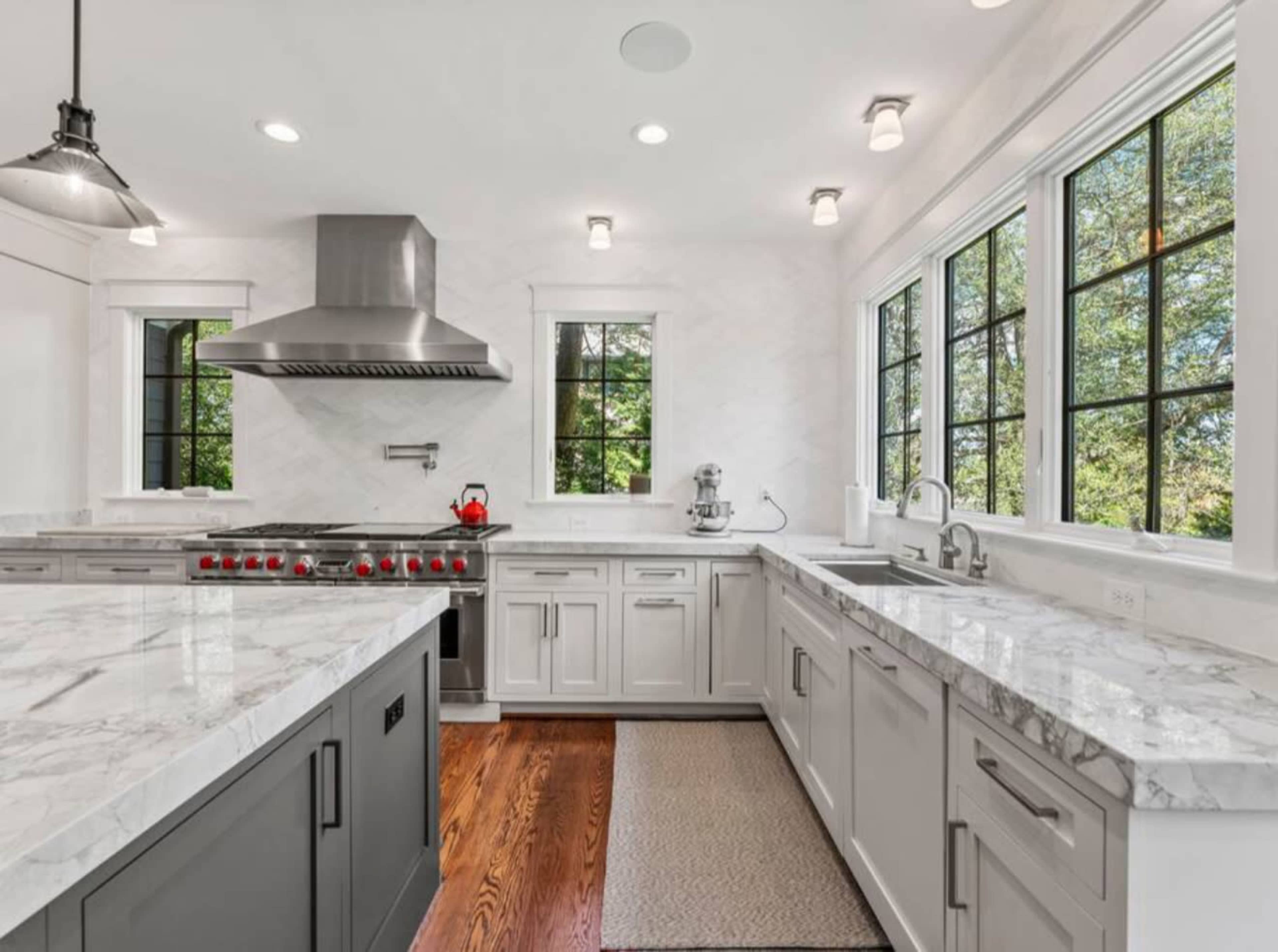 A modern kitchen features gray cabinets, a stainless steel range with red knobs, and large windows allowing natural light to illuminate the marble countertops.