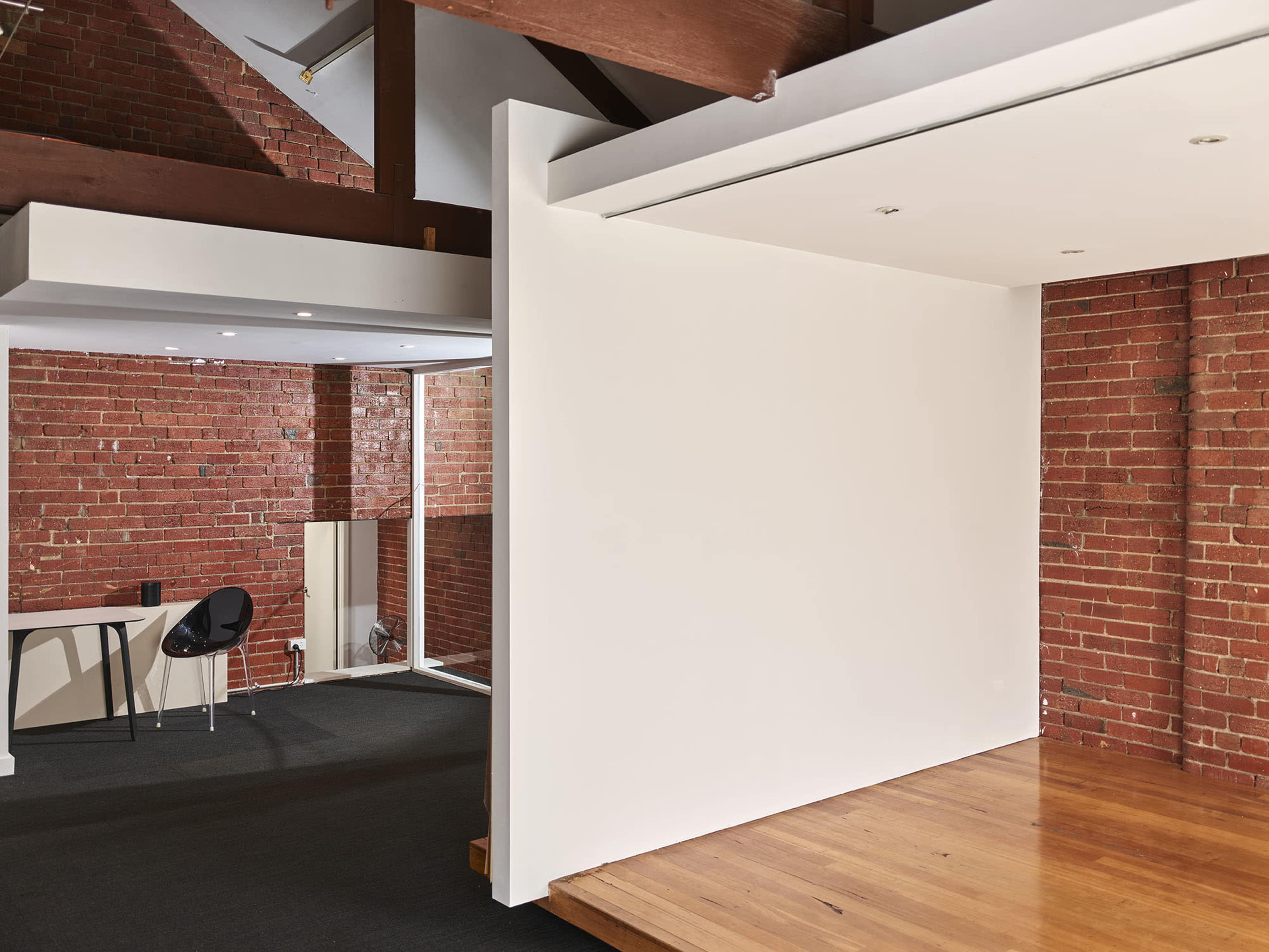 An interior space with exposed brick walls, wooden beams, a large blank wall, and a small desk with a chair.