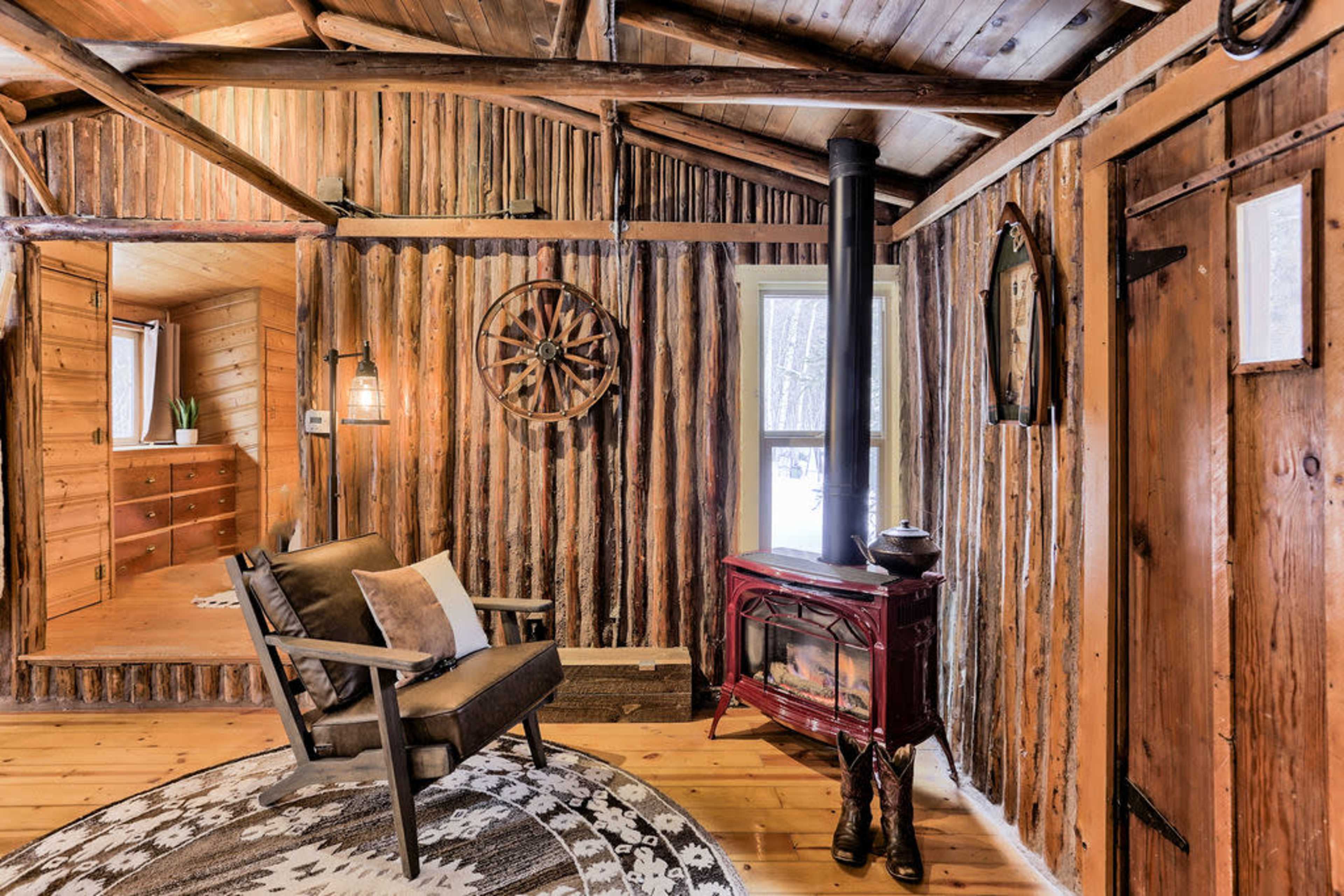 The interior of a rustic cabin features wooden walls, a stove, a chair, and a decorative wheel, with a small room visible in the background.