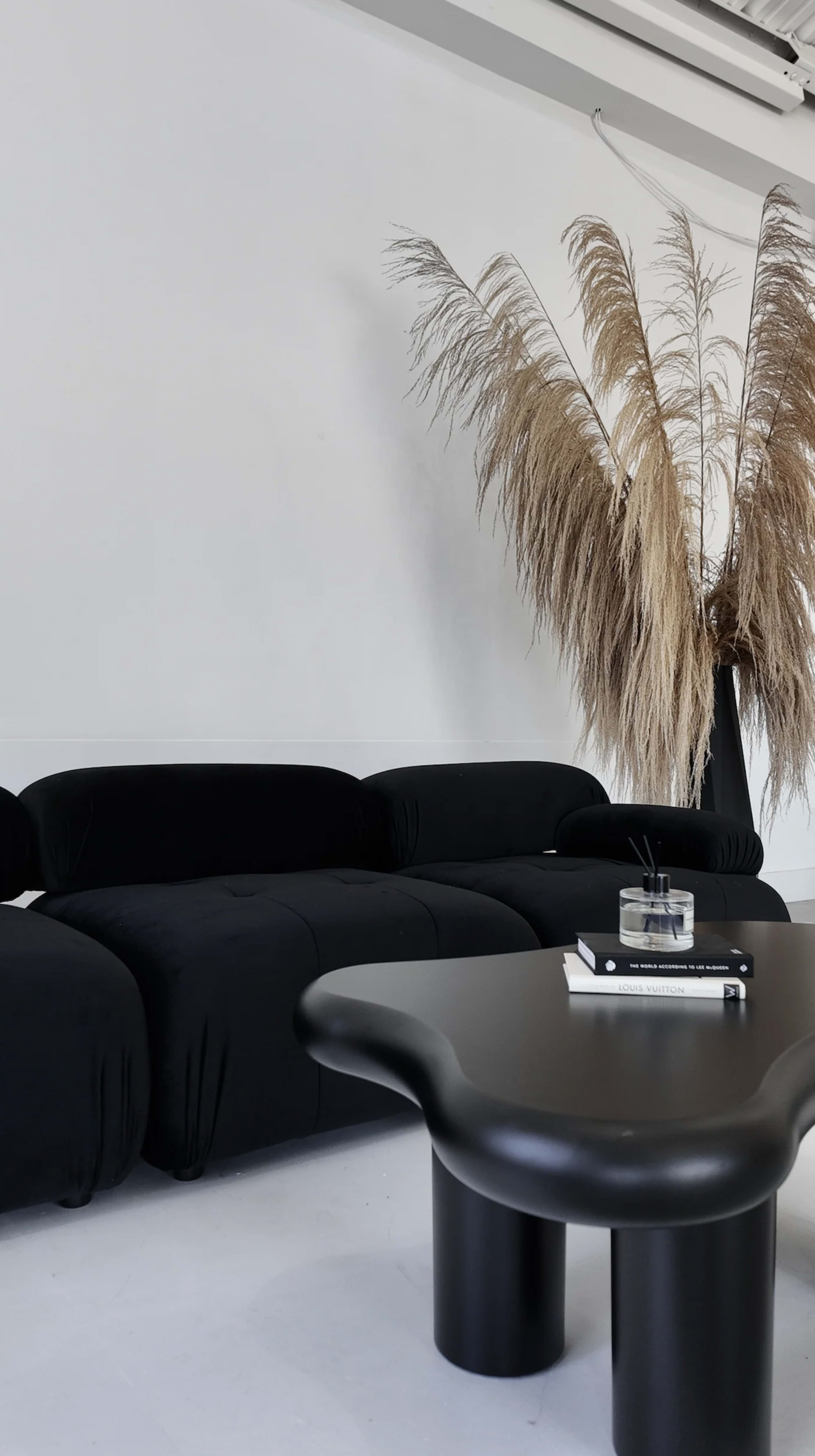 A black couch is positioned next to a large black vase containing dried pampas grass, with a low black table in front displaying a book and a candle.