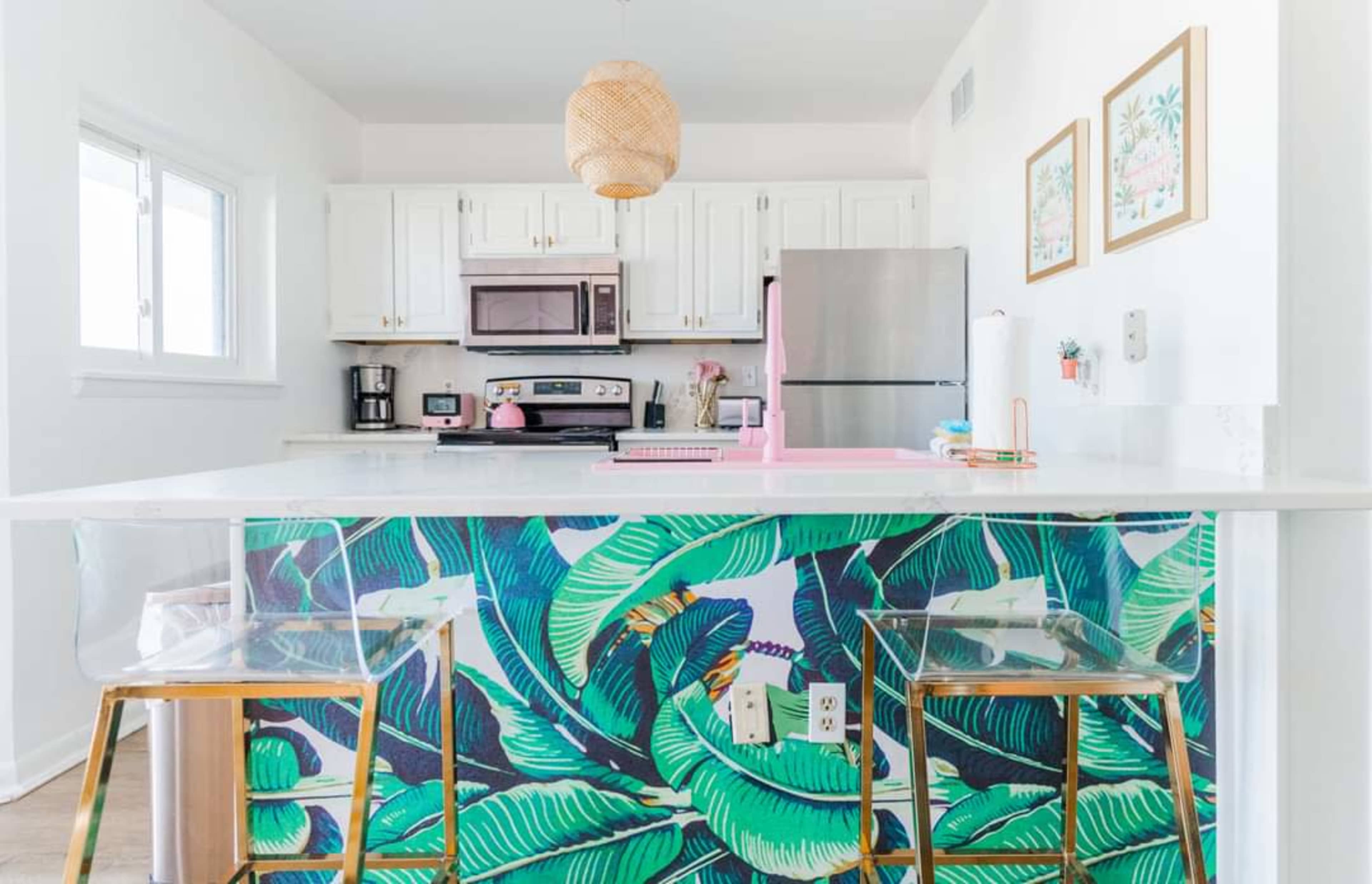 A modern kitchen features white cabinetry, stainless steel appliances, and a bar area with a tropical-themed backsplash and two clear chairs.