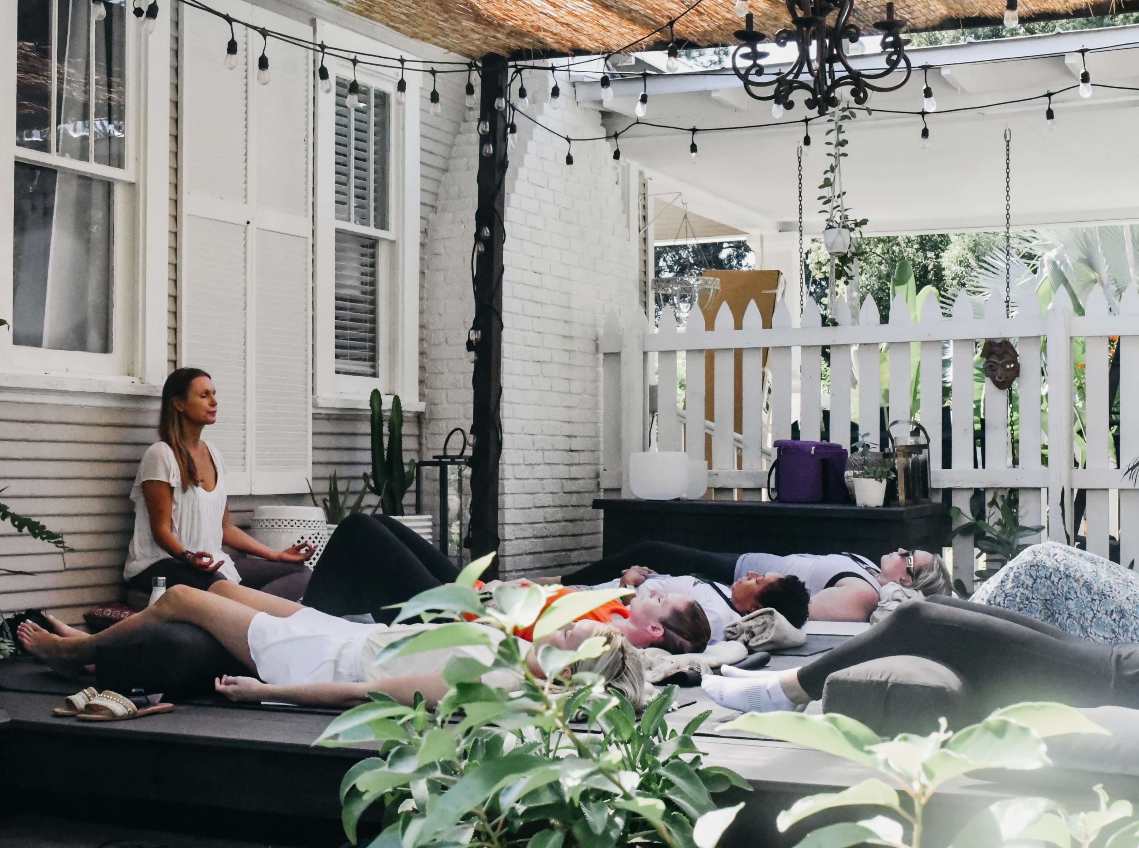A group of people relaxes on a shaded wooden deck, practicing mindfulness or meditation while seated on large cushions.