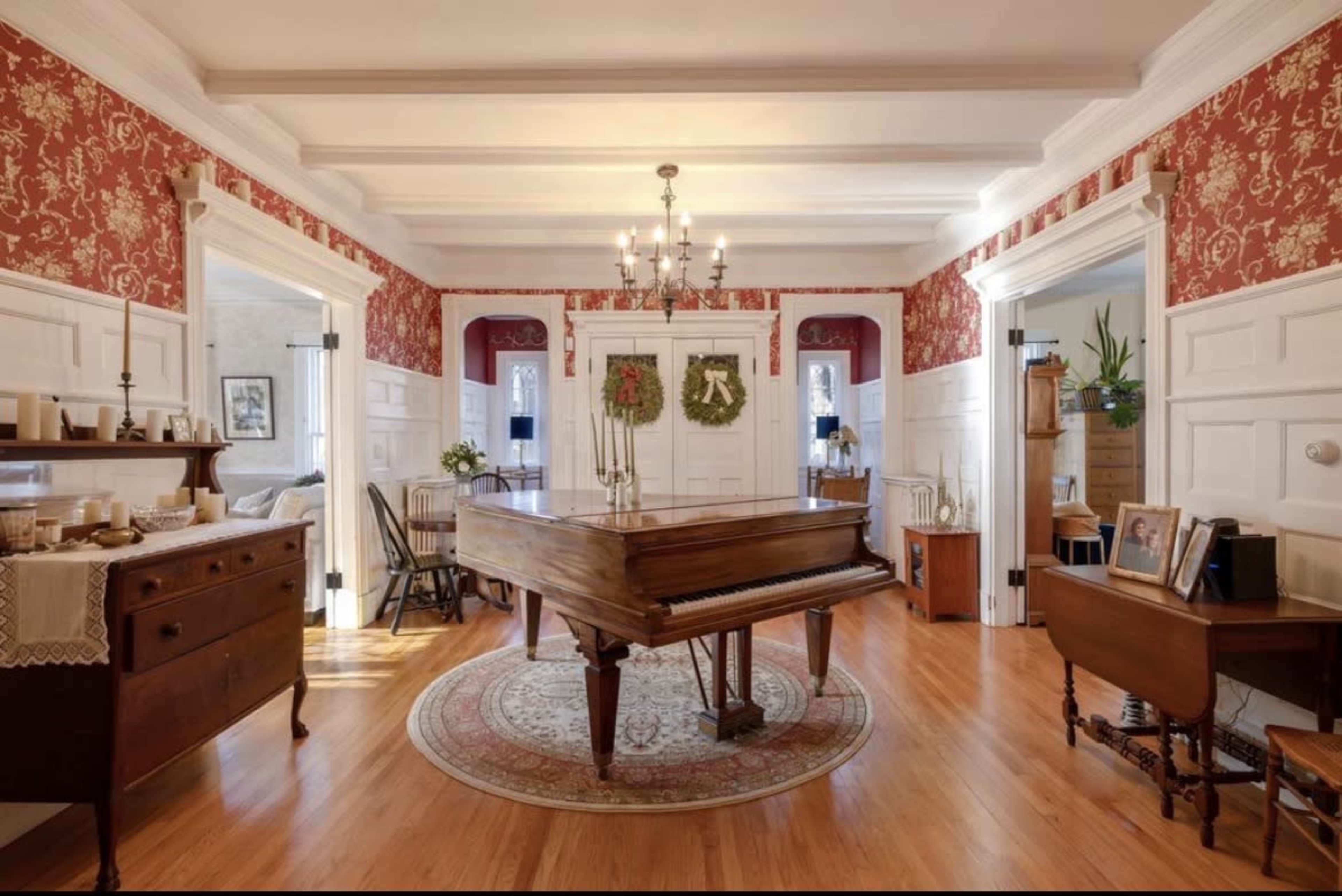 The image shows a warmly decorated room featuring a grand piano at its center, surrounded by patterned wallpaper and wooden flooring.