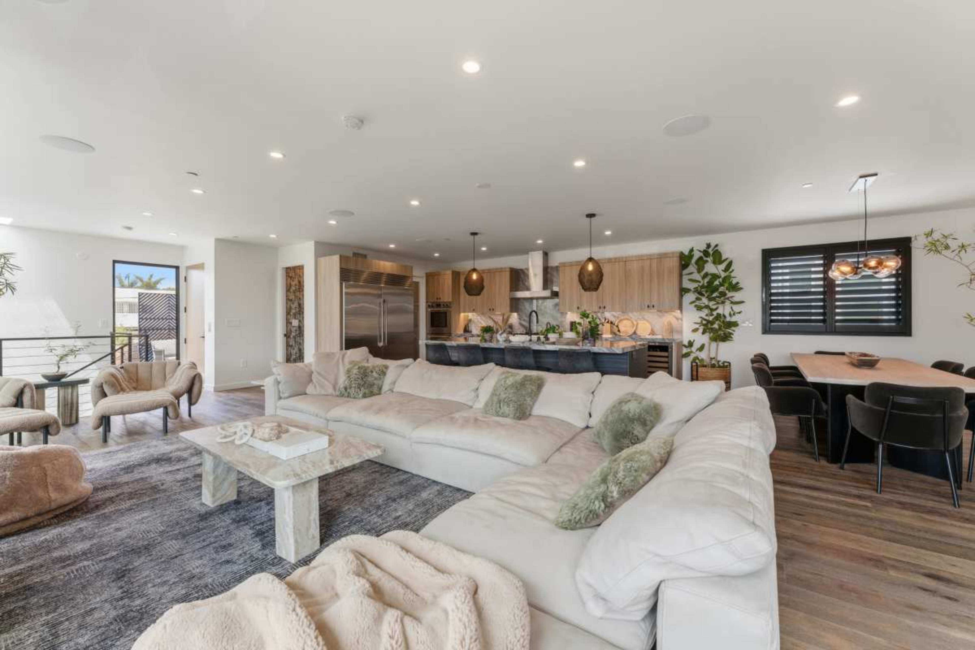 The image features a spacious modern living area with a large sectional sofa, a kitchen with sleek cabinetry, and a dining table set for several occupants.