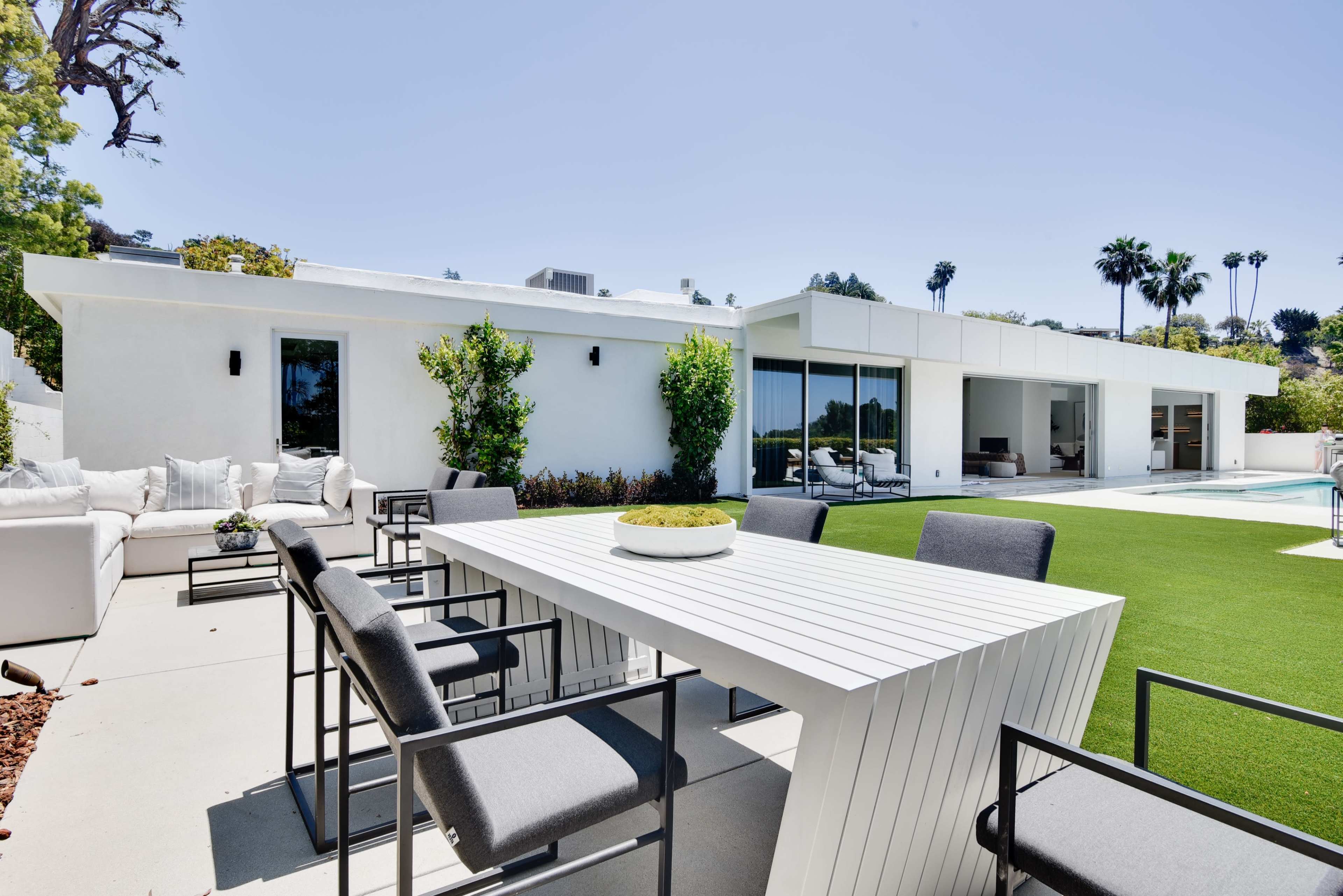 A modern outdoor dining area features a white table surrounded by gray chairs, adjacent to a swimming pool and a neatly maintained lawn.