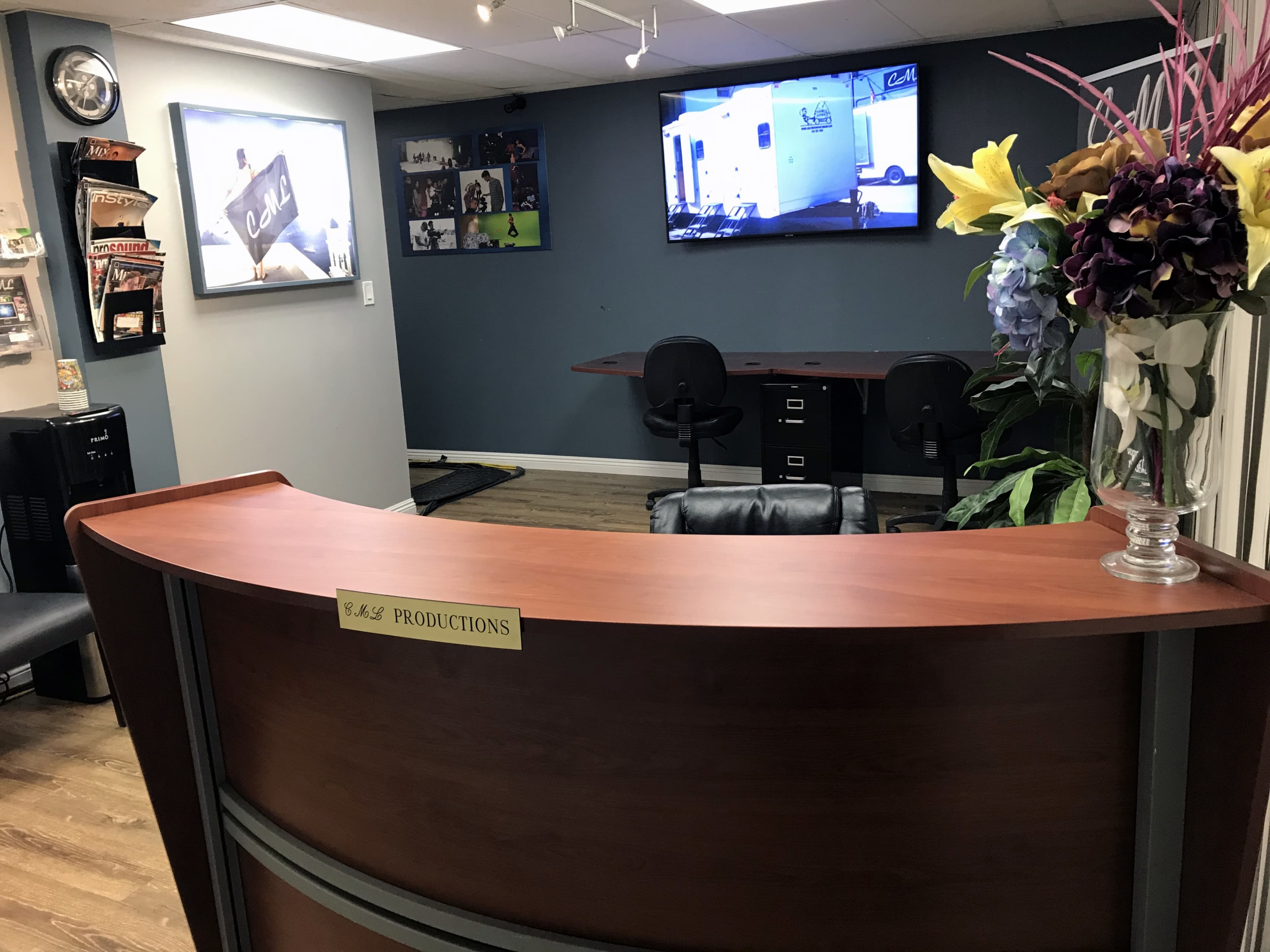 A reception area with a curved wooden desk labeled "Productions," a television mounted on the wall, and a workspace in the background.