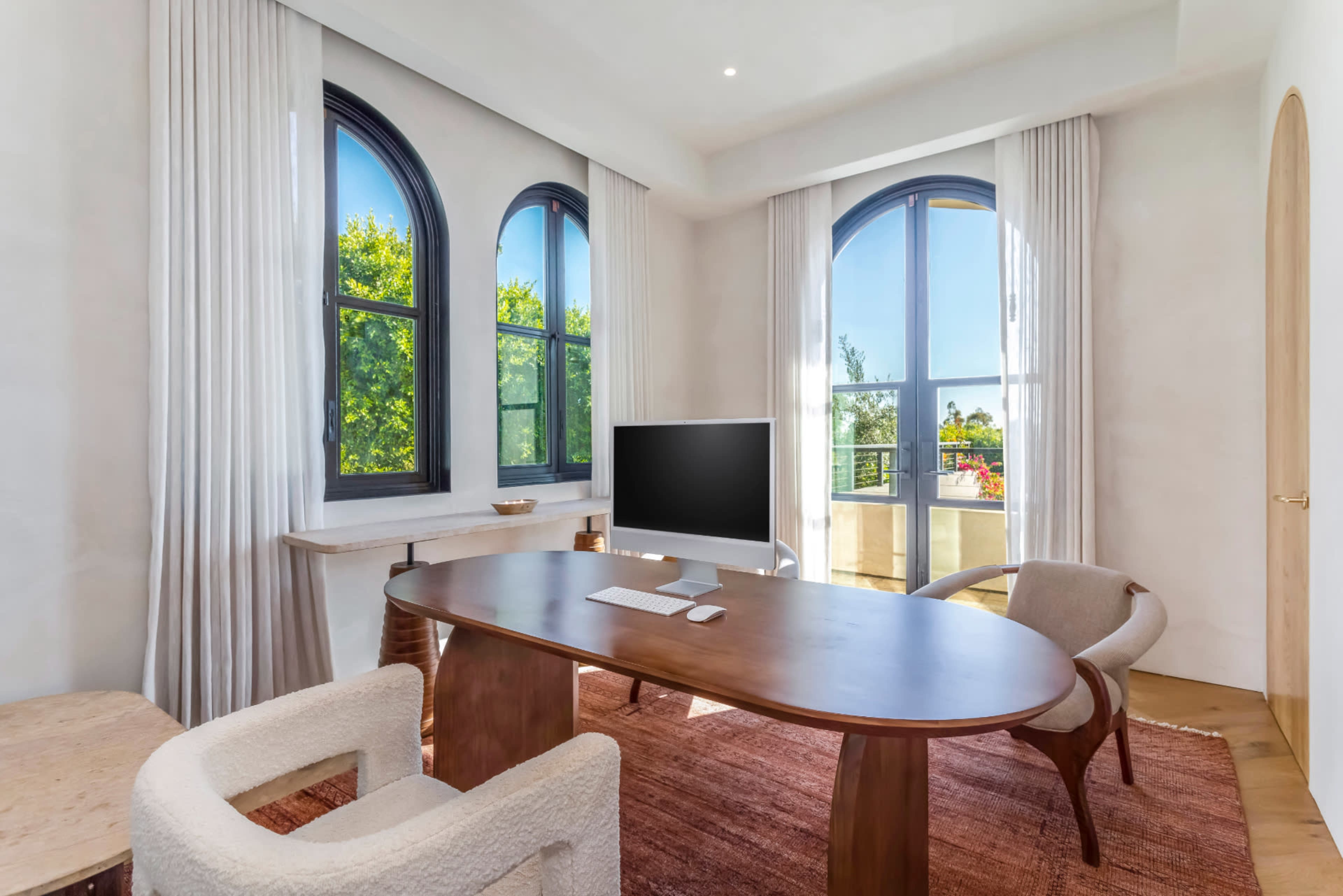 A modern office features a large wooden desk, a computer monitor, and windows with arched frames, letting in natural light.
