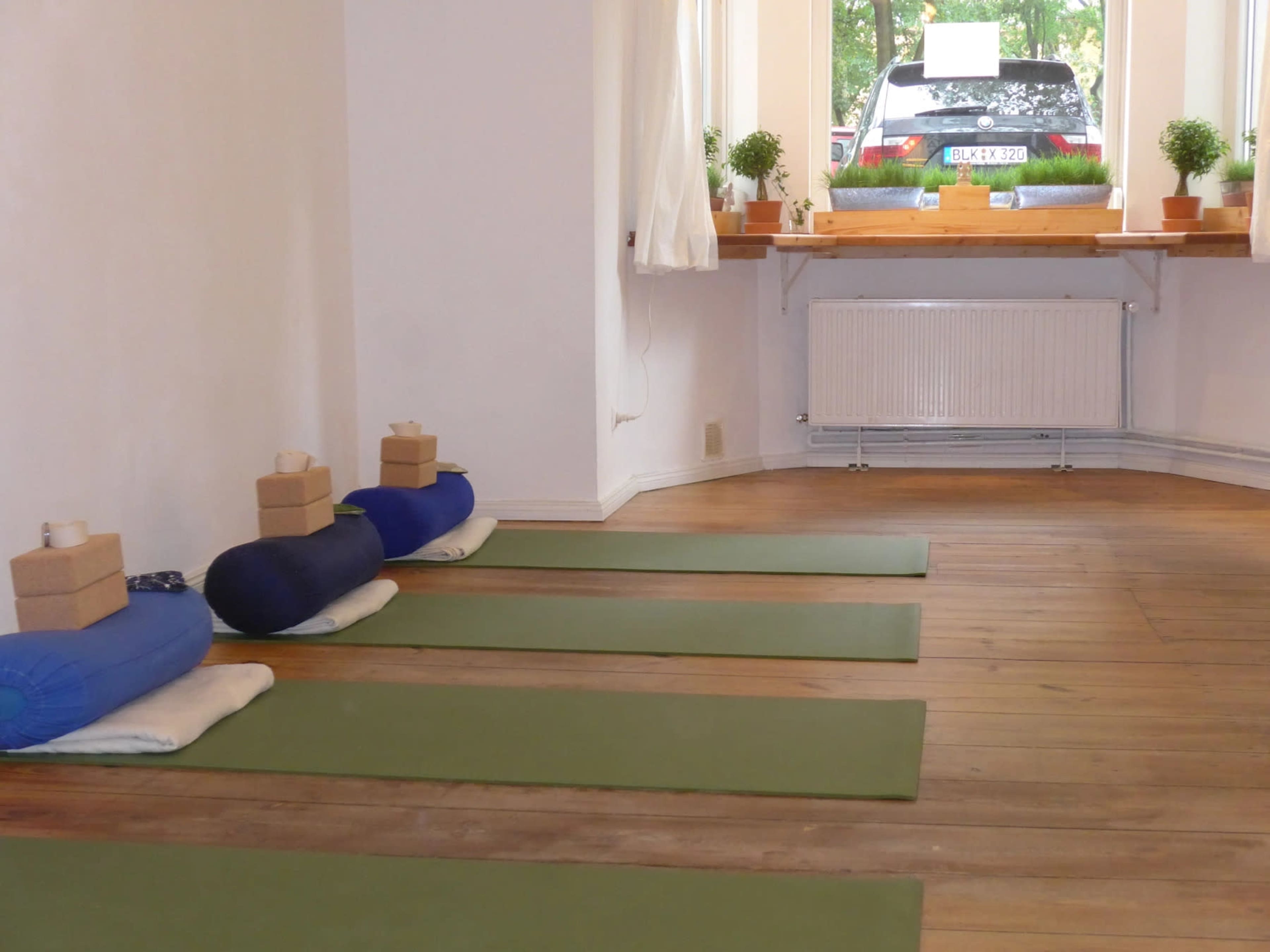 The image shows a yoga studio with green mats arranged on a wooden floor, blocks placed on top of rolled mats, and a car visible through a window.
