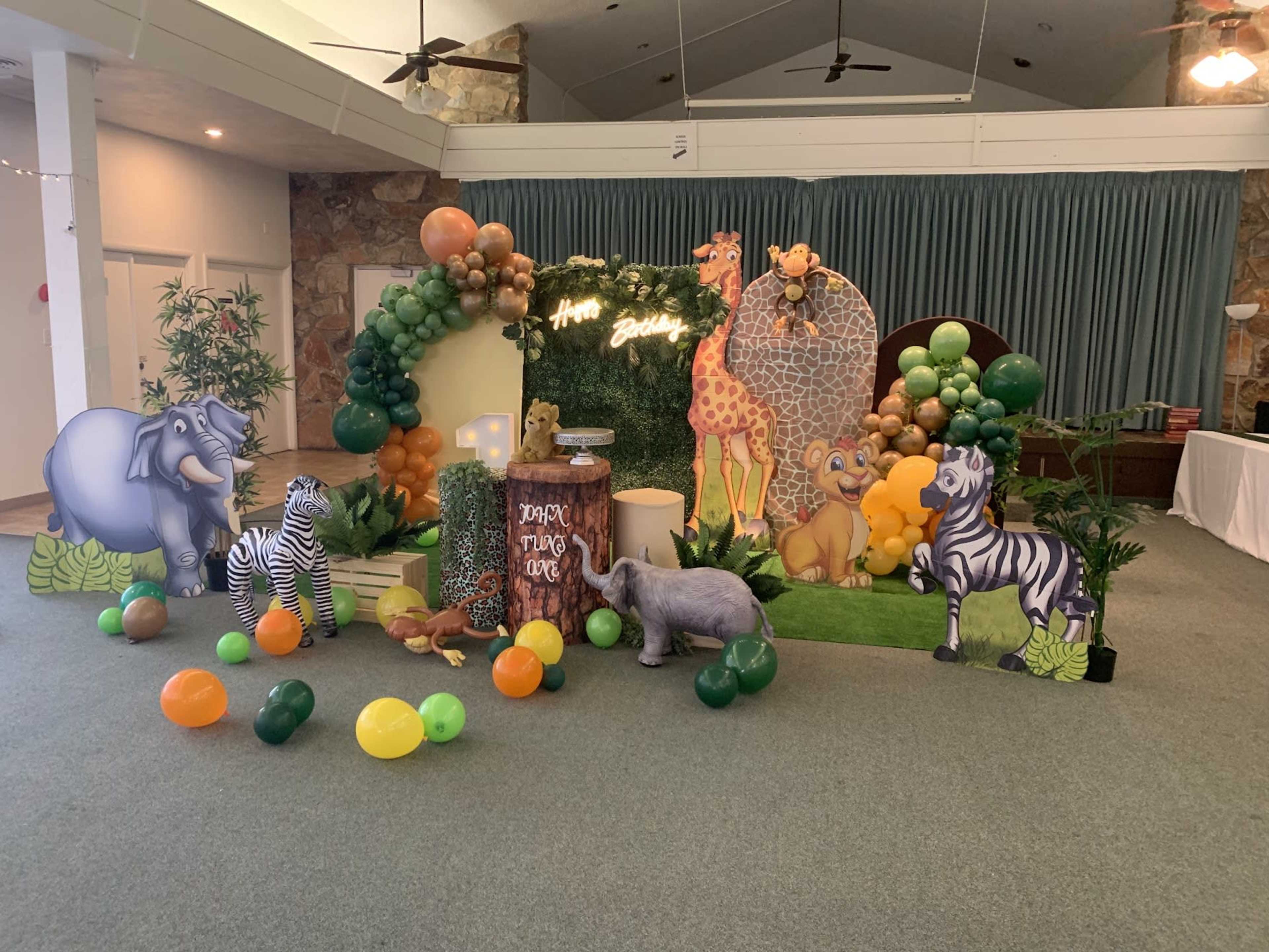 The scene features a vibrant jungle-themed decorations setup with animal cutouts, balloons, and a greenery backdrop for a celebration.