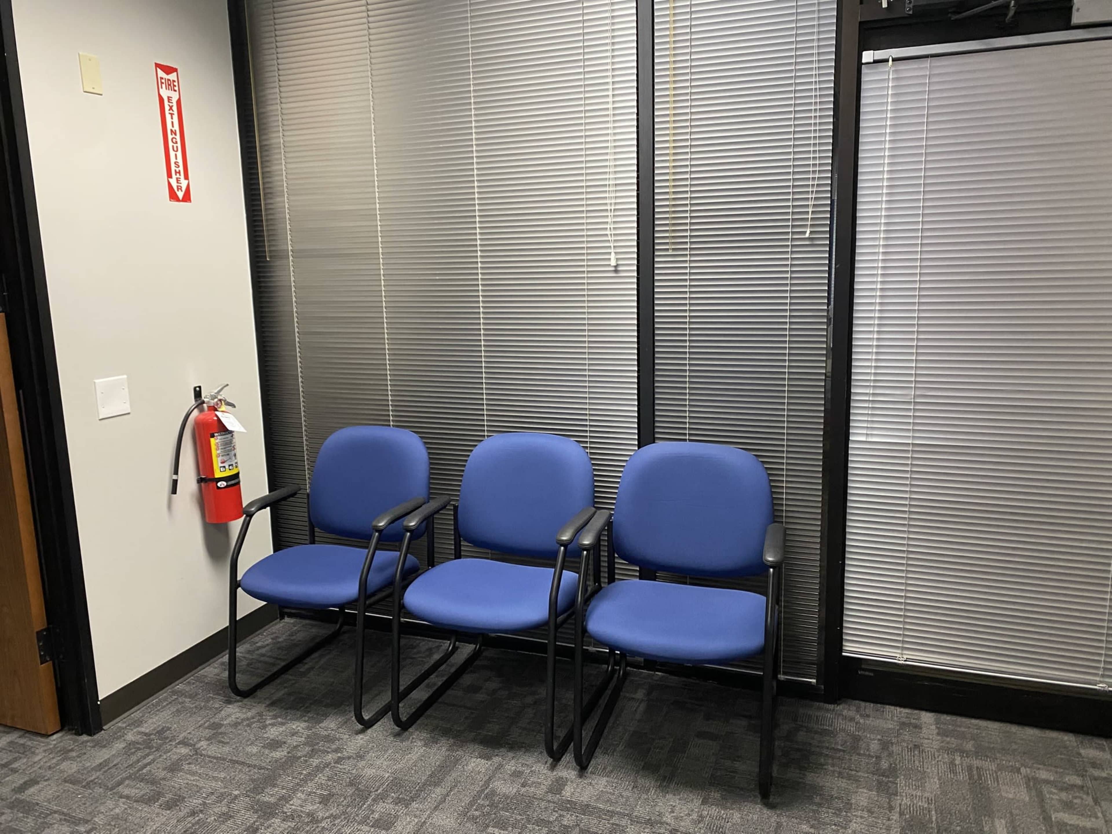 The image displays three blue chairs arranged in a row against a wall with blinds, alongside a fire extinguisher.