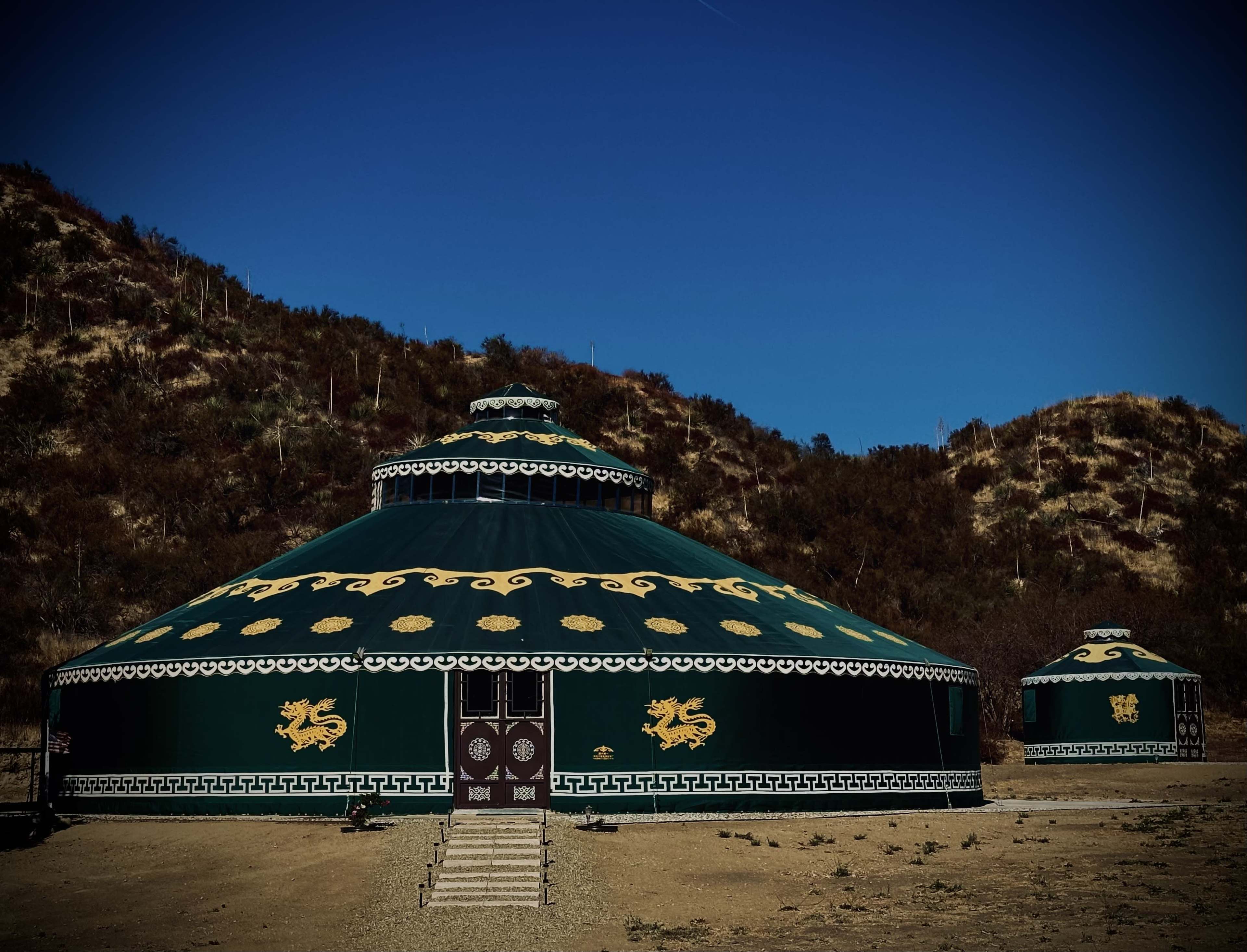 Two large green yurts with ornate designs stand against a backdrop of rolling hills under a clear blue sky.