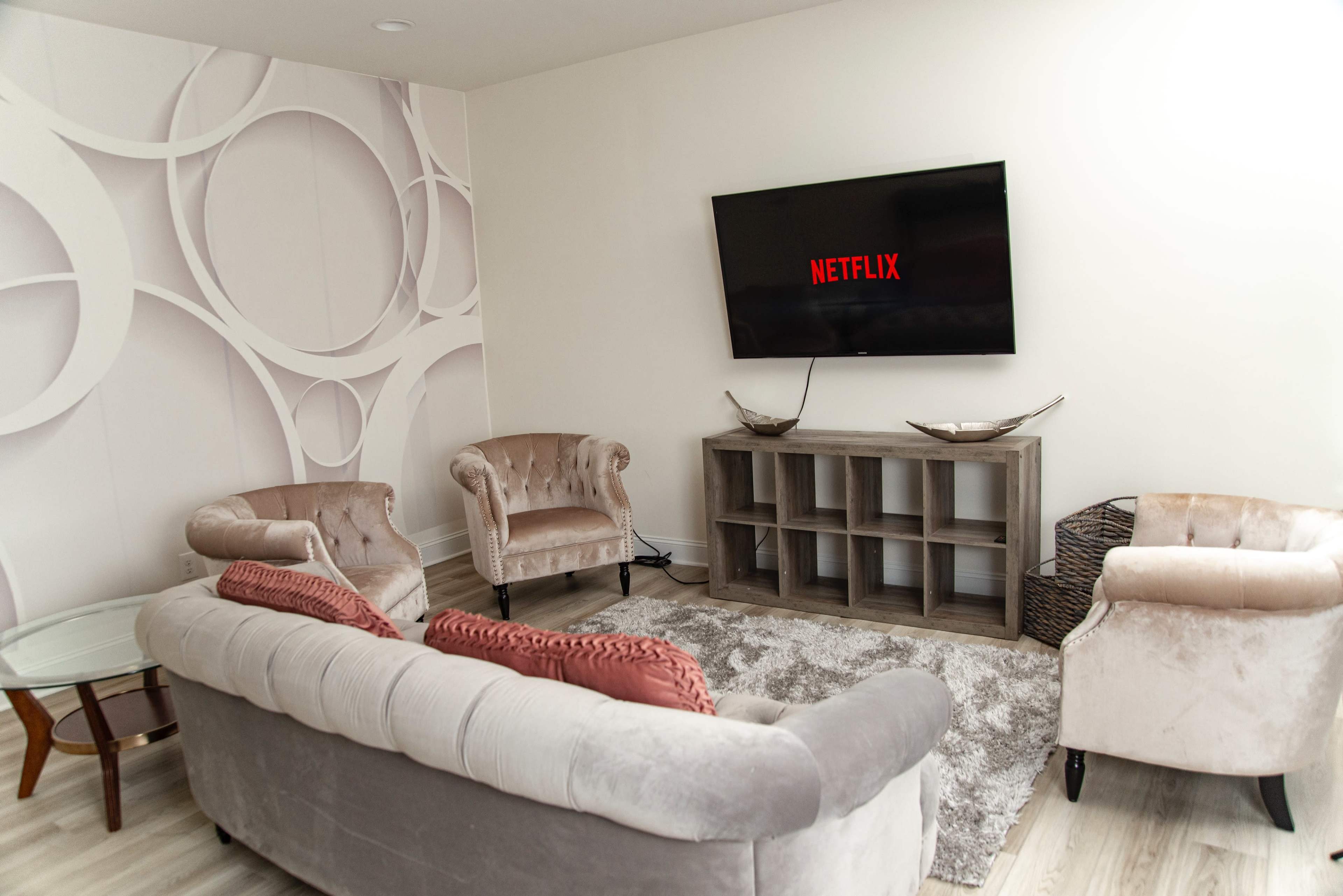 A modern living room features a large television displaying the Netflix logo, flanked by two elegant armchairs and a gray sofa, with a coffee table and small shelf unit.