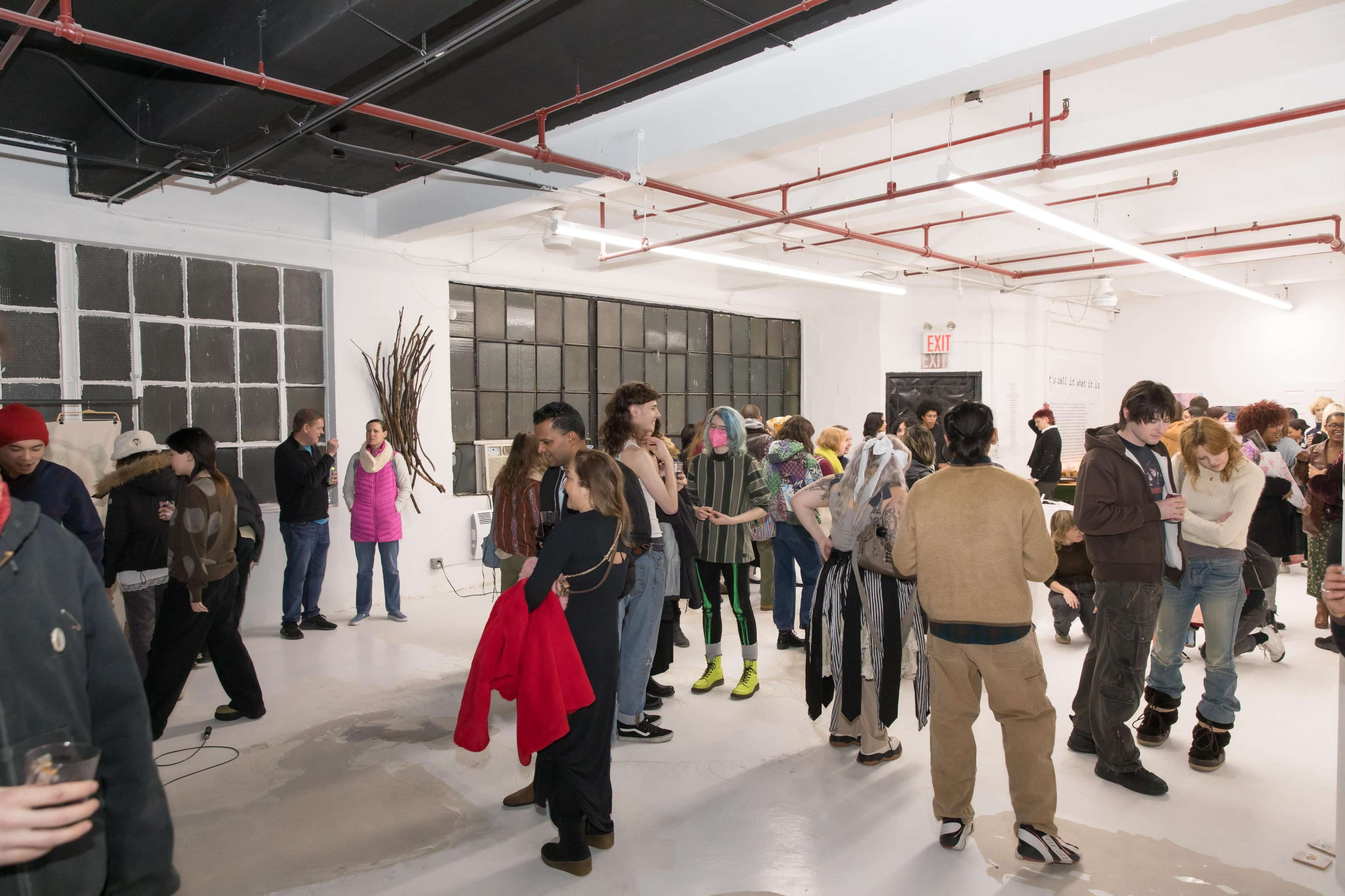 A group of people is gathered in a spacious, modern art gallery with industrial-style elements, engaging in conversation and socializing at an event.