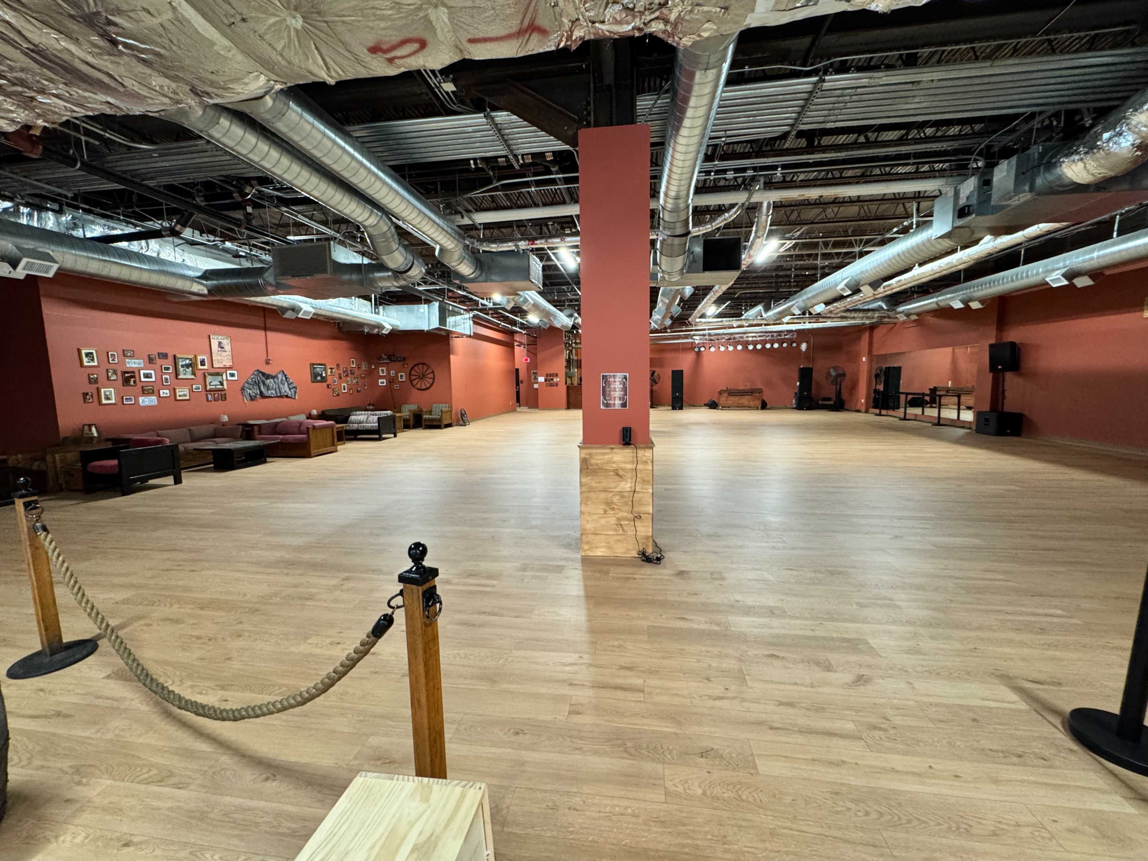 The image shows an empty interior space with wooden flooring, red walls, and industrial-style air ducts overhead, featuring minimal furniture and sound equipment.