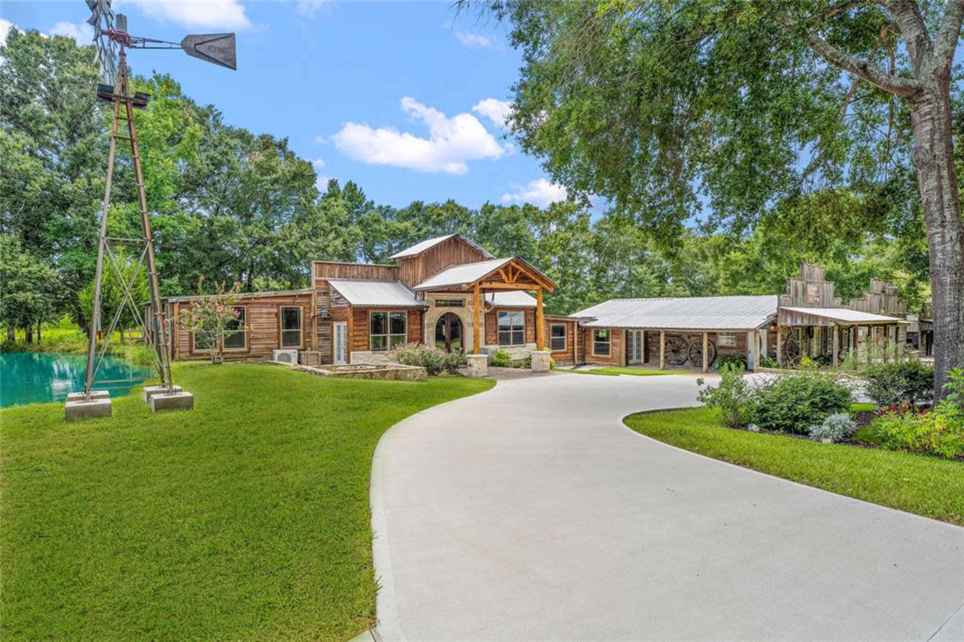 A rustic-style house surrounded by greenery, with a circular driveway leading to the entrance and a windmill nearby.