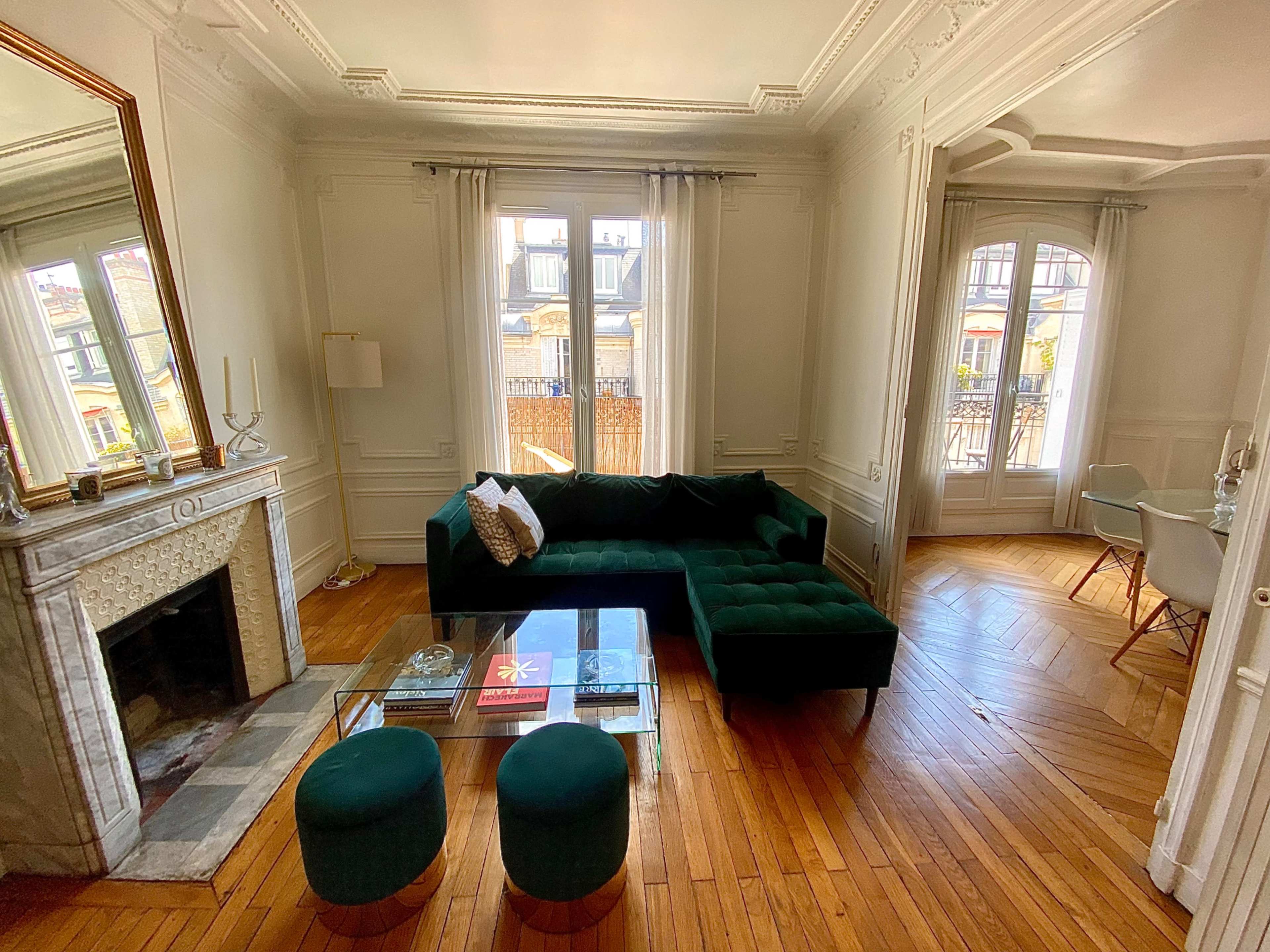 A stylish living room featuring a green velvet sectional sofa, a glass coffee table, and large windows allowing natural light to enter the space.