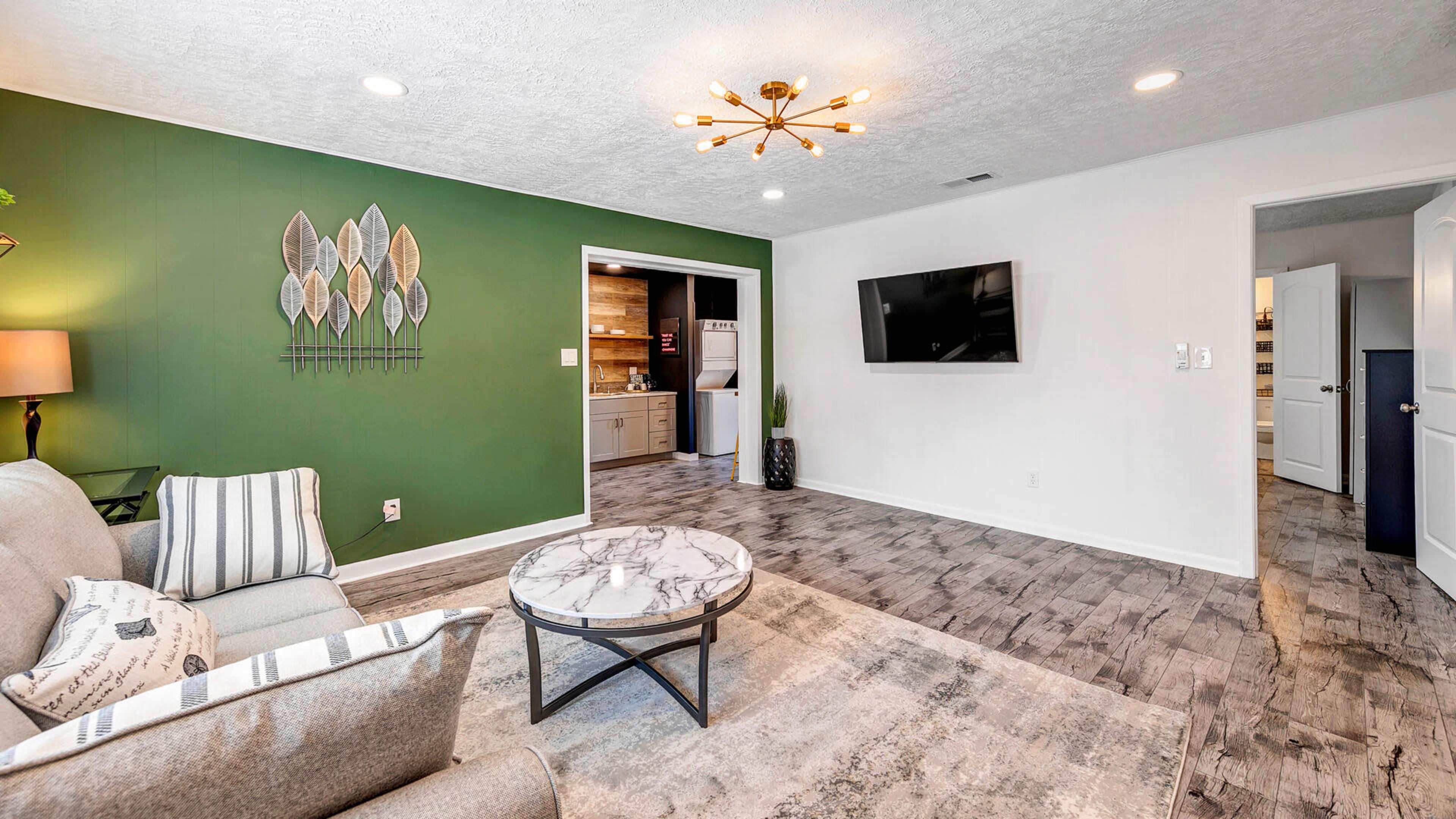A living room features a green accent wall, a gray sofa, a round marble coffee table, and a television mounted on the wall.