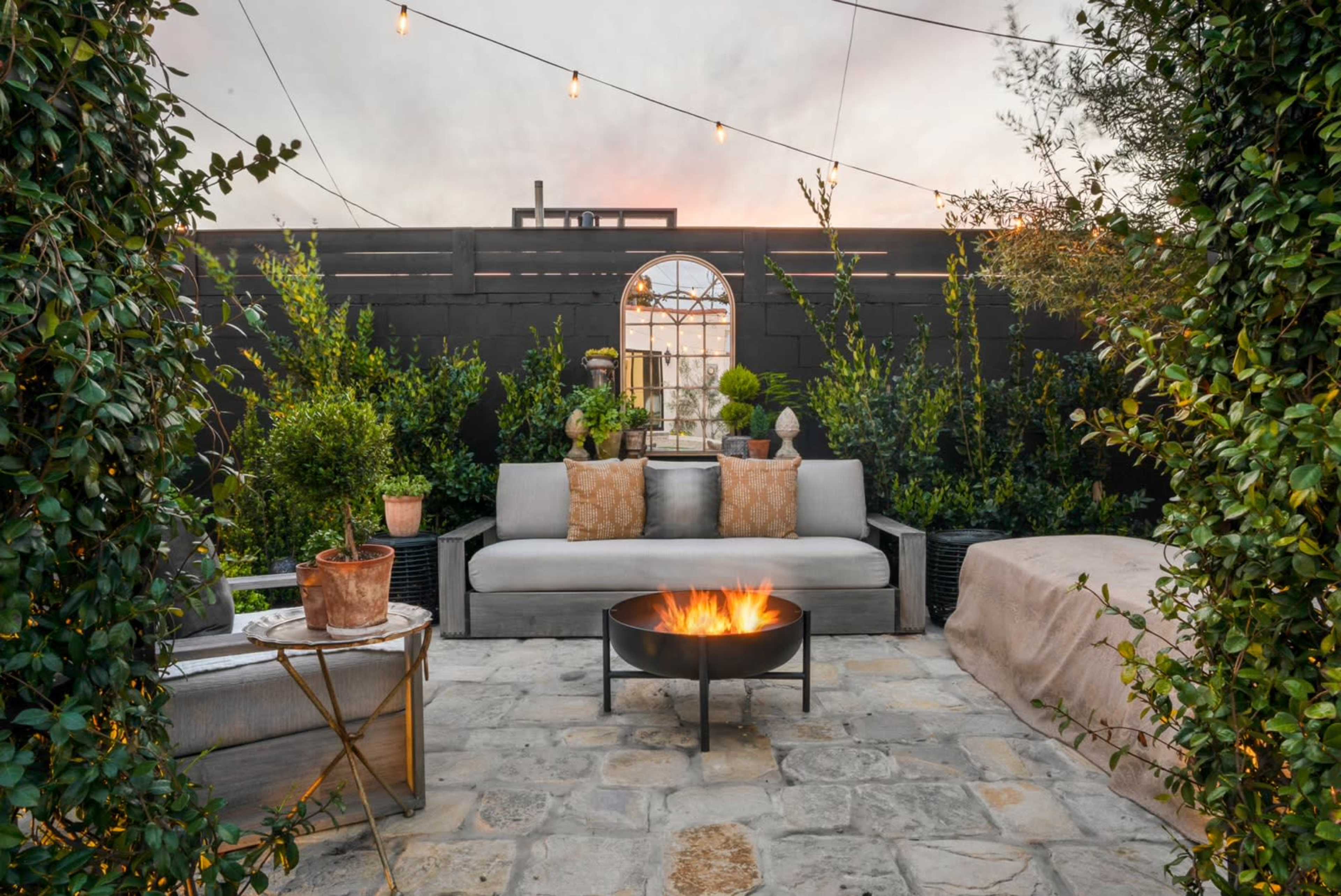 A cozy outdoor seating area with a fire pit, surrounded by plants and decorative elements under a string of lights.