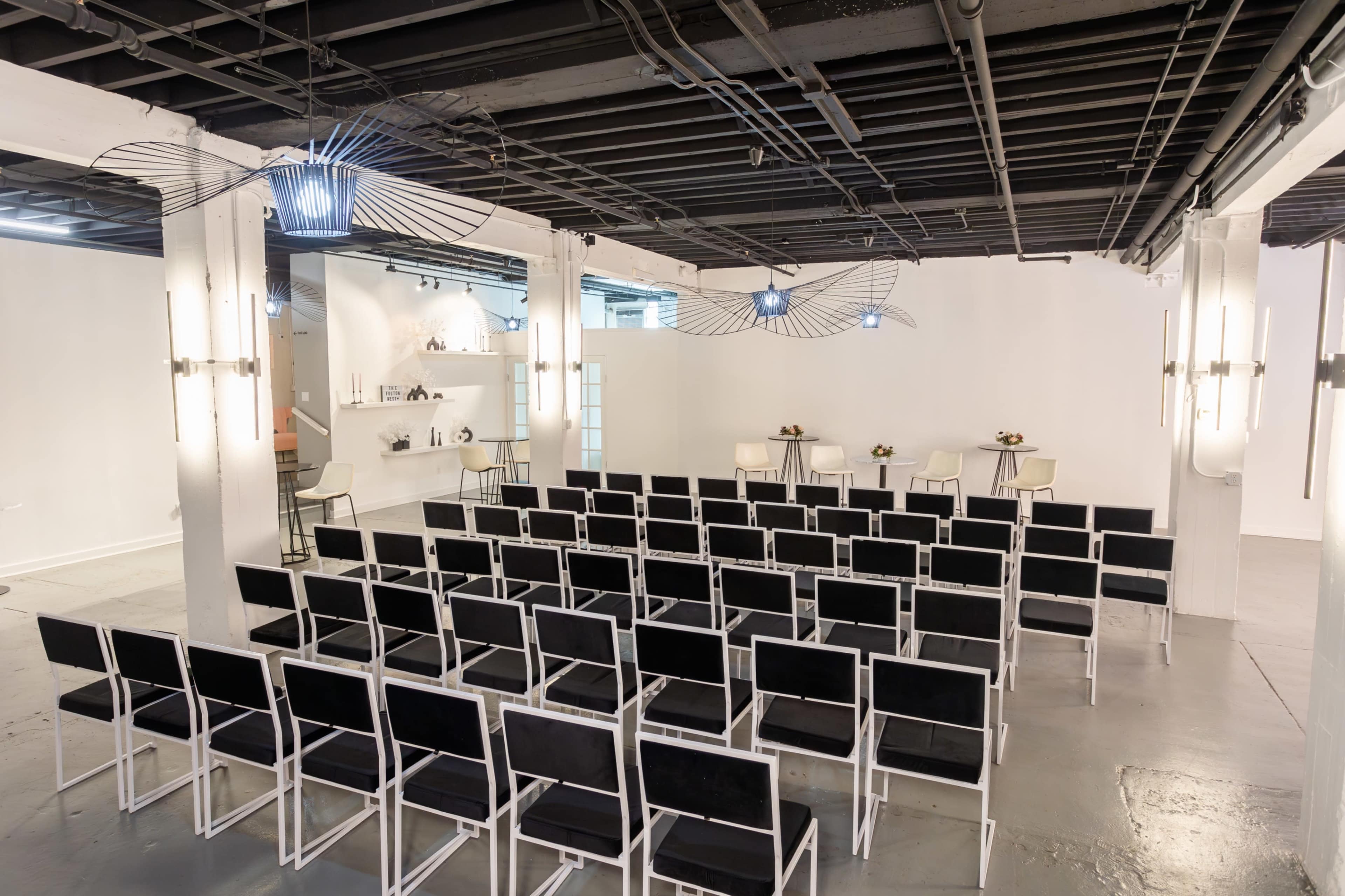 A spacious room features rows of black chairs arranged facing a minimalistic counter with two stools, illuminated by modern lighting fixtures.