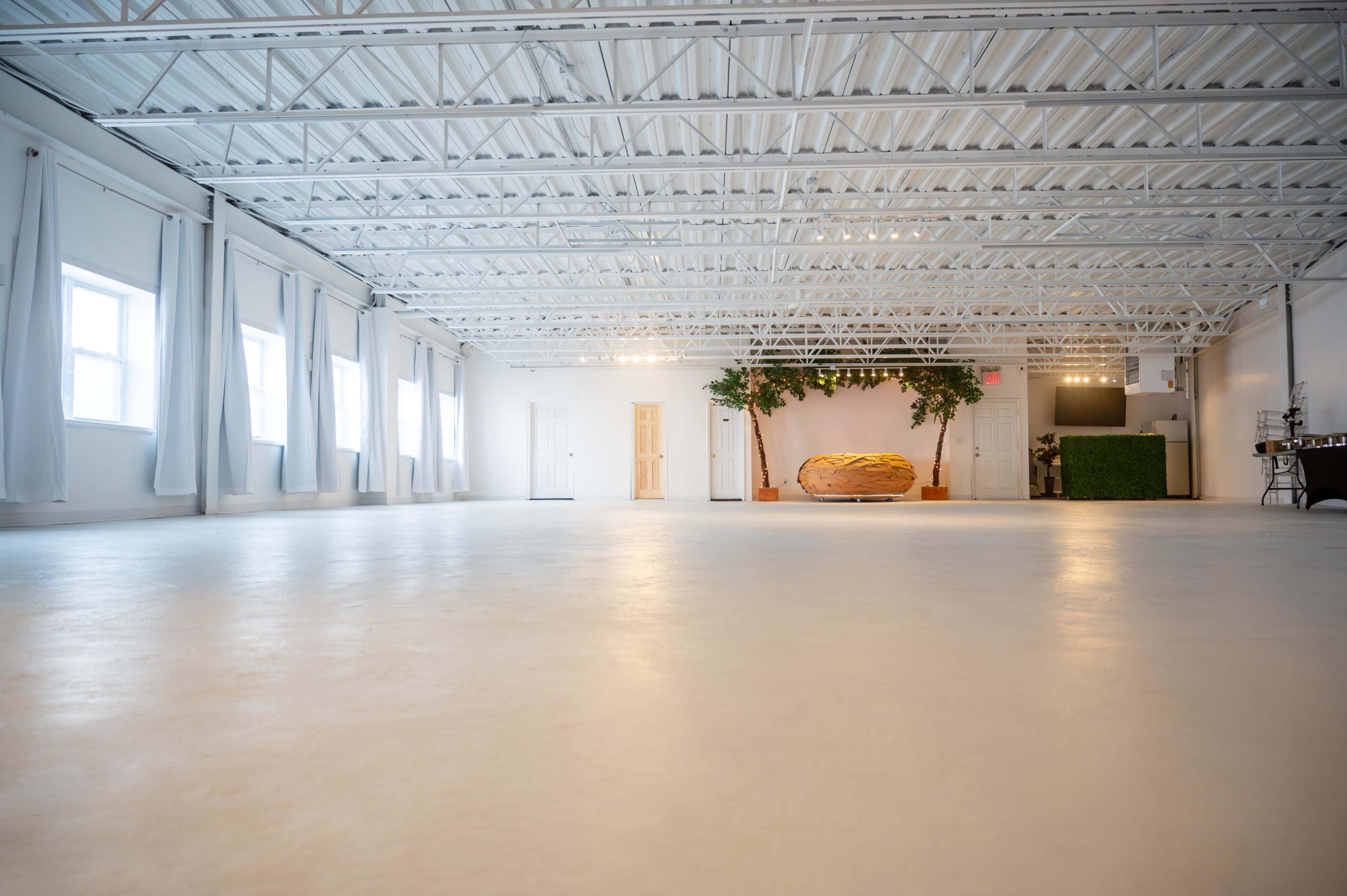 The image shows a spacious, well-lit interior with white walls, a smooth floor, and minimalistic decor, featuring a large, yellow structure in the center and greenery in the background.