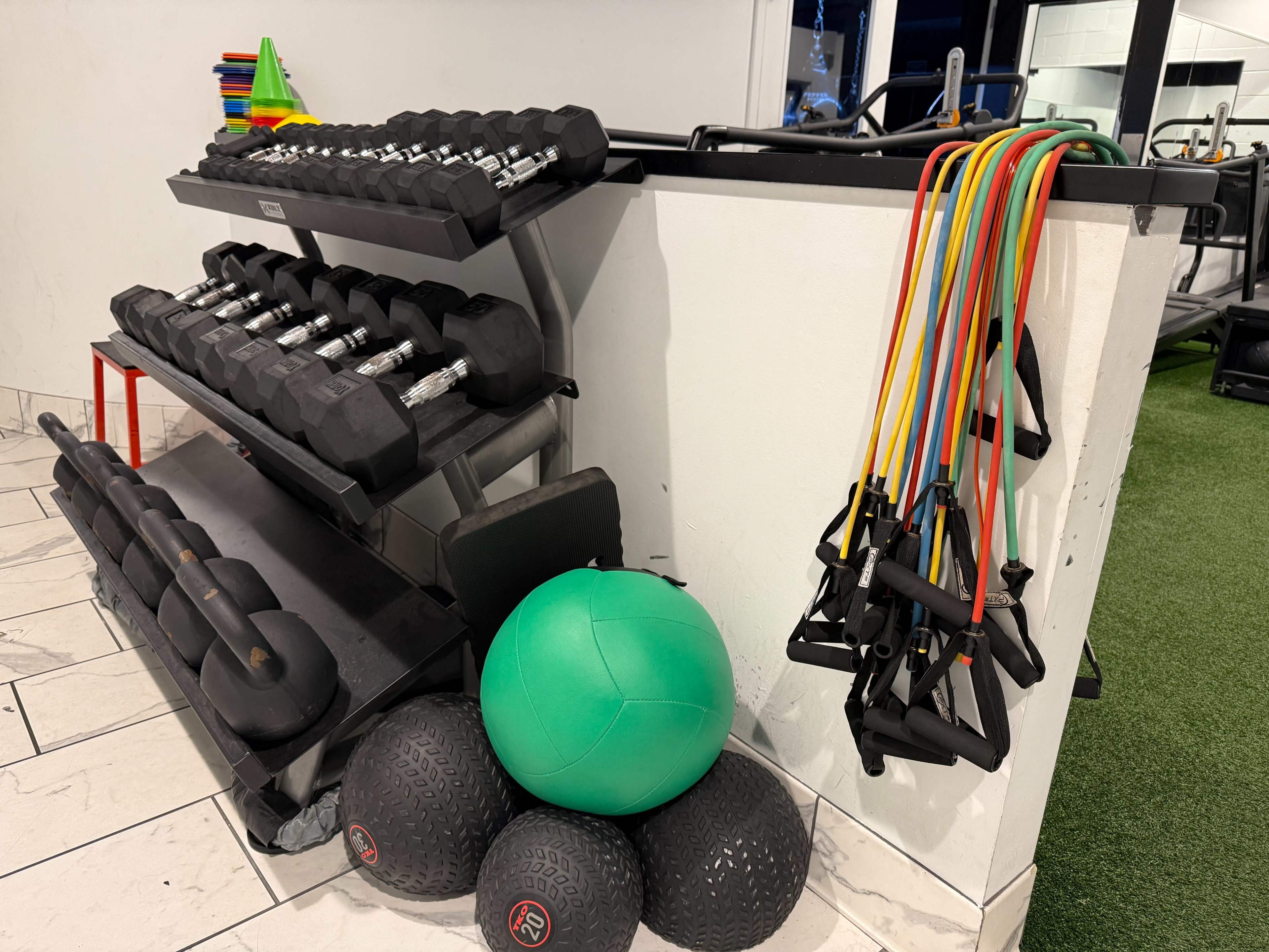 The image shows a gym area featuring a rack of dumbbells, resistance bands hanging on a wall, and several exercise balls arranged on the floor.