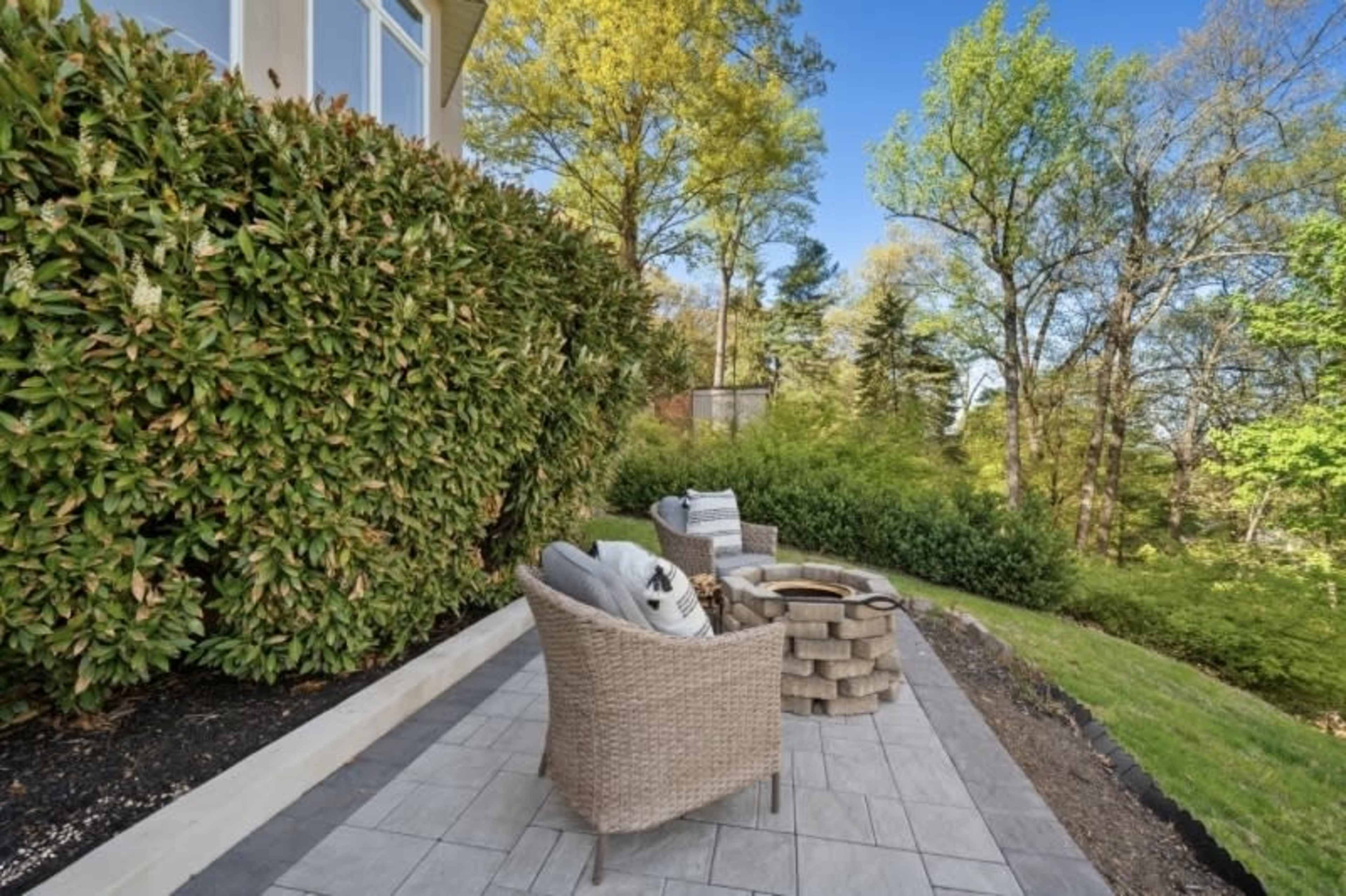 A patio area features wicker chairs and a stone fire pit, surrounded by lush greenery and trees.