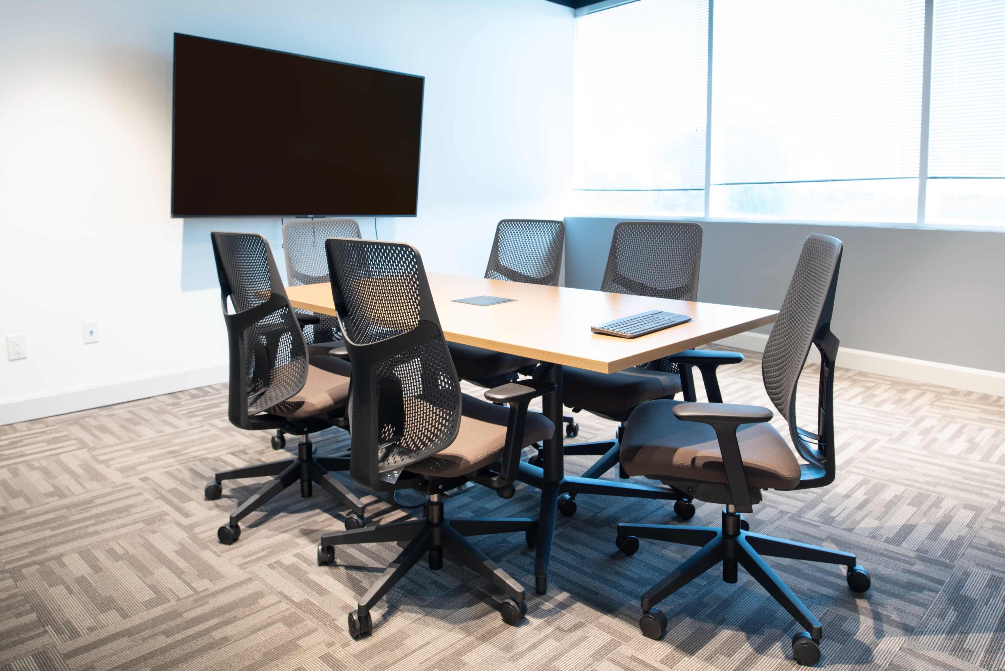 A conference room is arranged with a rectangular table surrounded by six ergonomic chairs, and a large screen mounted on the wall.