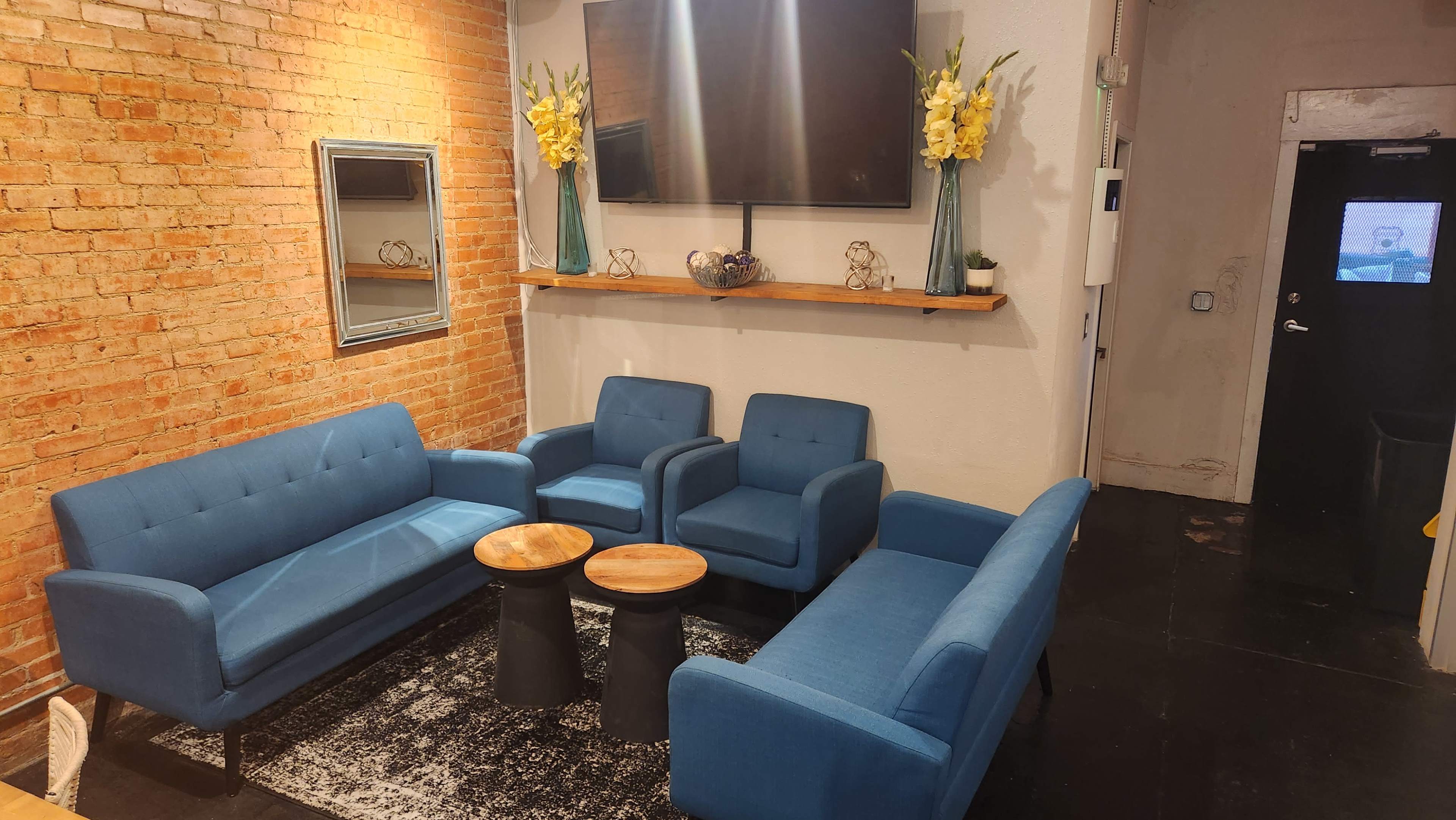 A small seating area with blue sofas and armchairs arranged around two round tables on a patterned rug, against a backdrop of exposed brick walls and a wall-mounted TV.