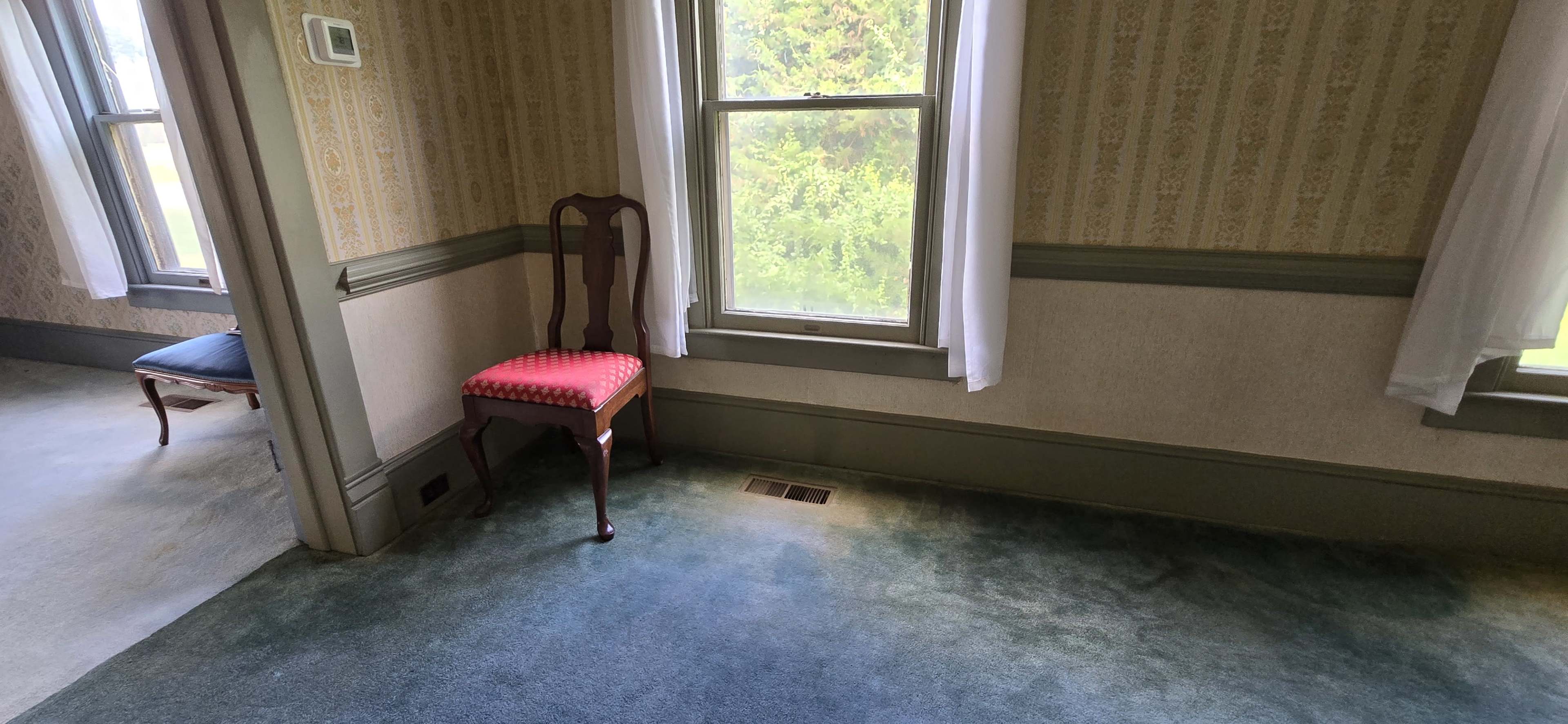 The image shows a room with beige patterned wallpaper, a green carpet, large windows with white curtains, a wooden chair with a red padded seat, and a blue upholstered chair in the background.