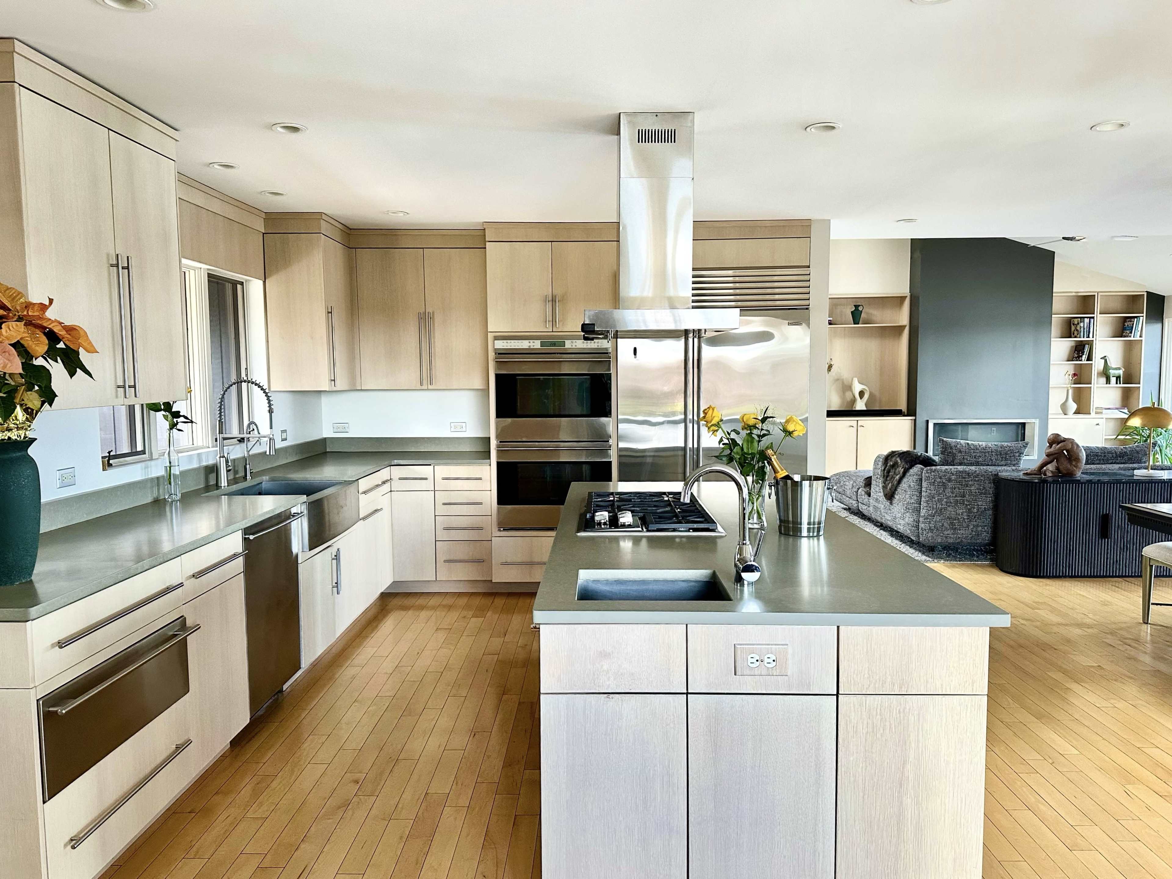 The image shows a modern kitchen featuring light wood cabinetry, stainless steel appliances, and a central island with a sink, surrounded by an open living area.