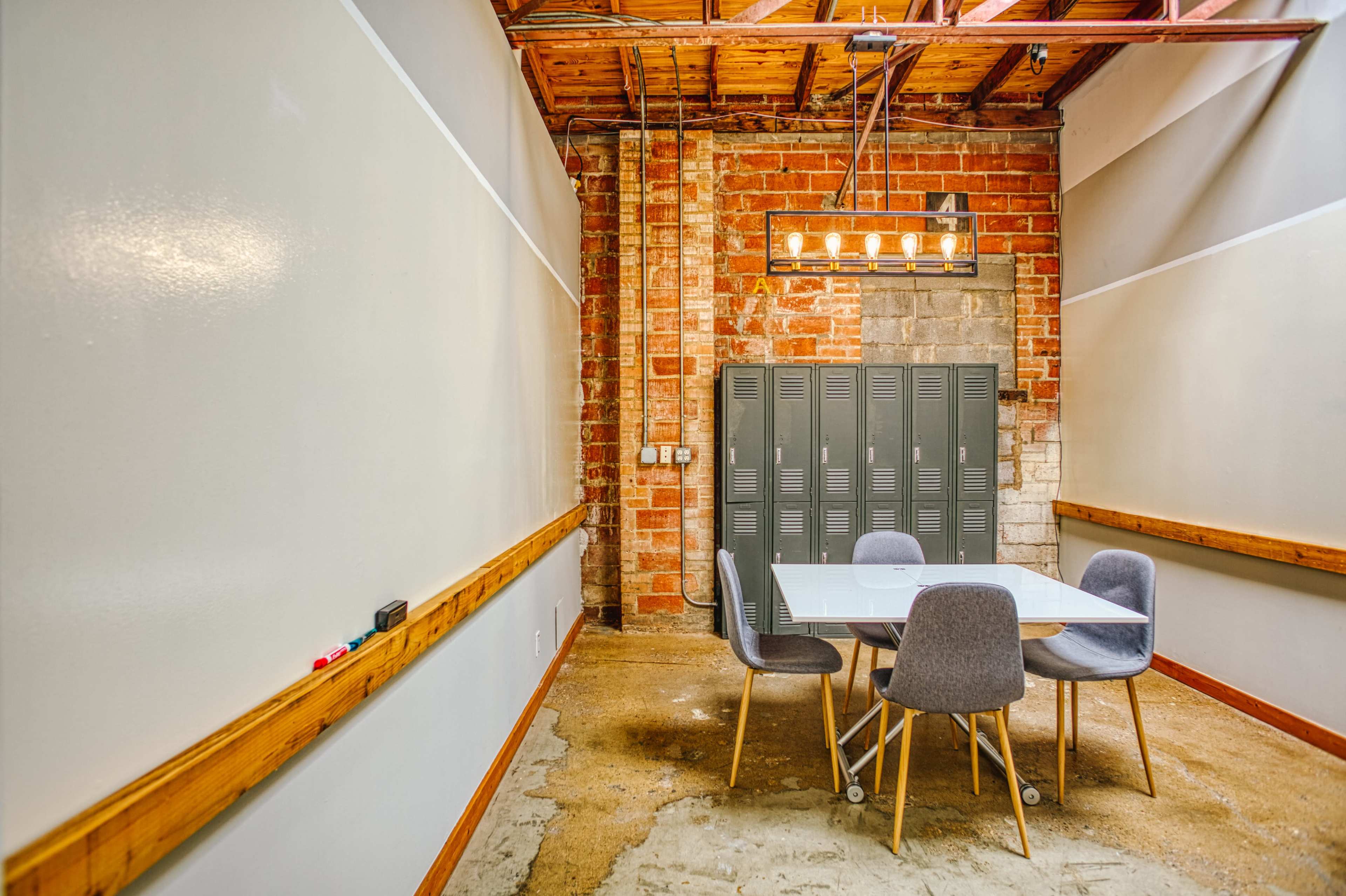 A conference room features a glass table surrounded by four gray chairs, with exposed brick walls and metal lockers in the background.