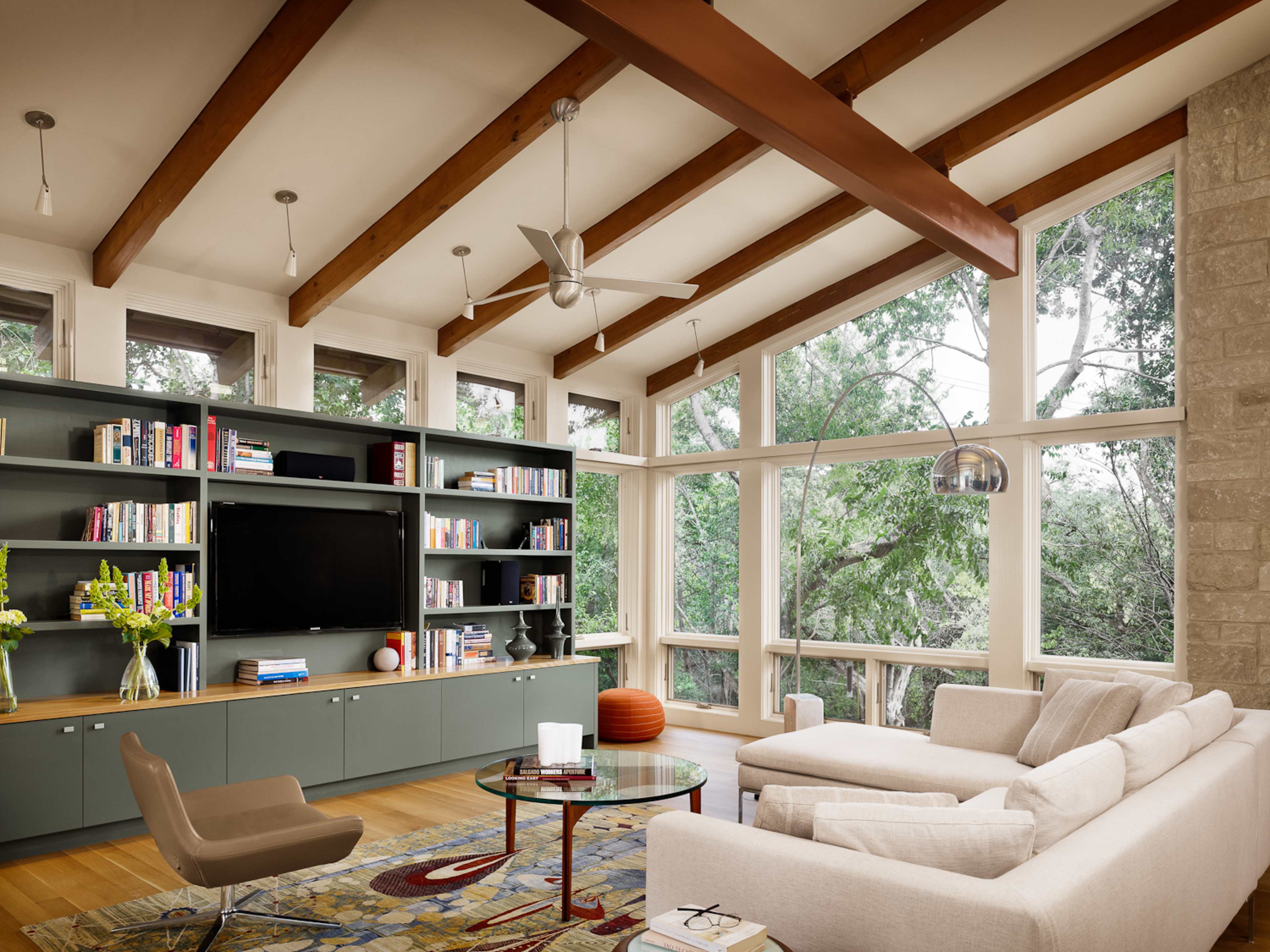 A modern living room with a beige sectional sofa, a glass coffee table, a large bookshelf, and windows overlooking greenery.