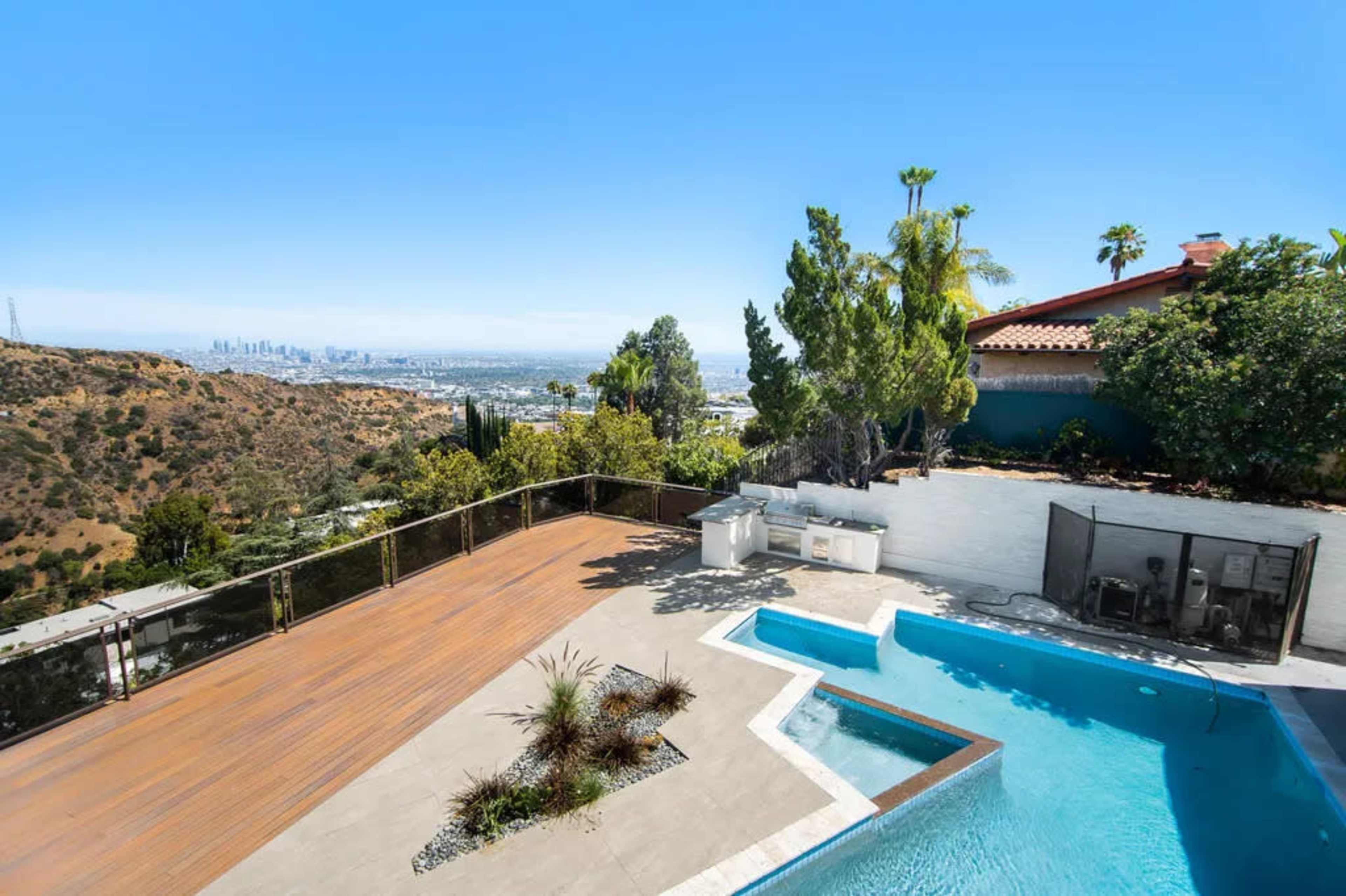 The image shows a hillside property featuring a swimming pool, wooden deck, and scenic views of a city skyline in the background.