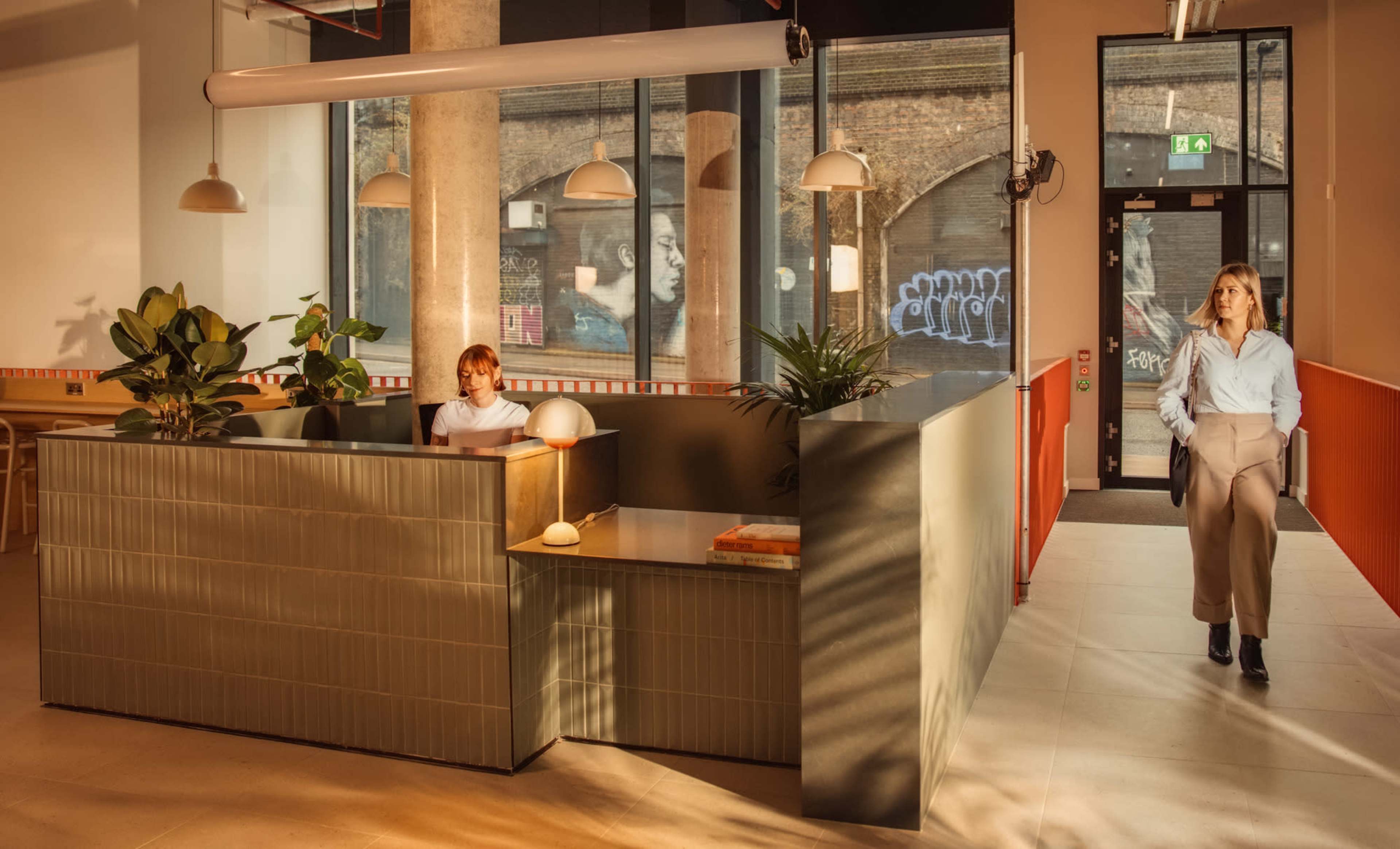 A modern office reception area features a woman at a front desk with plants nearby, while another woman walks in through the entrance illuminated by natural light.