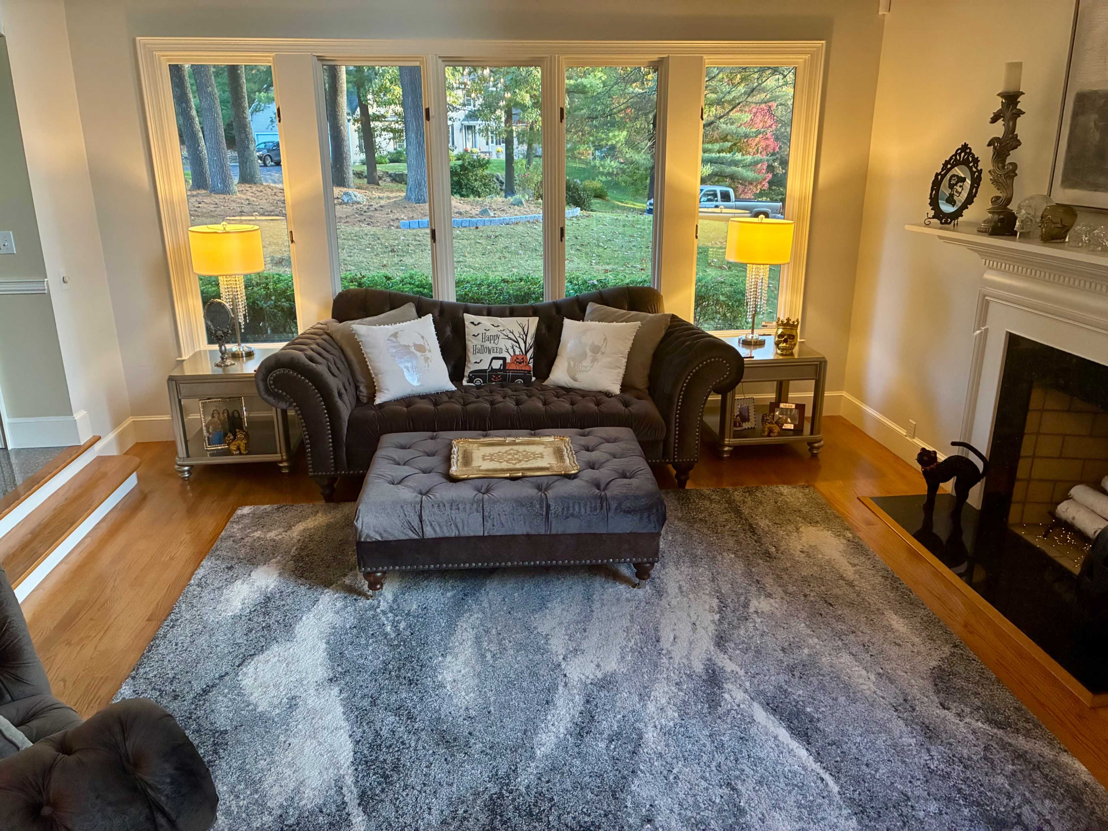 The living room features a dark sectional sofa and an ottoman on a large area rug, with bay windows allowing natural light and framed by two table lamps.