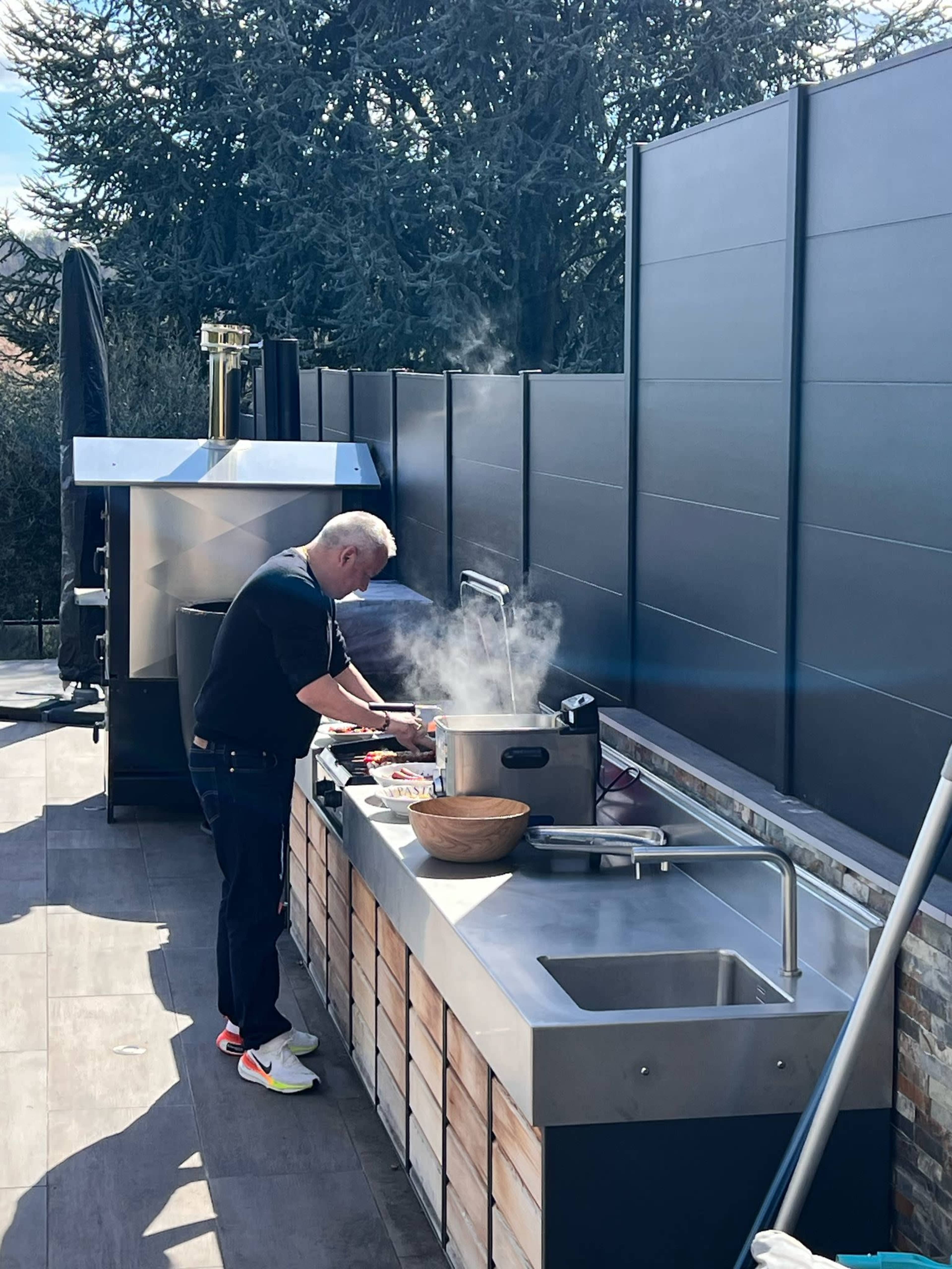 A person is cooking at an outdoor kitchen, with steam rising from a cooking pot, on a sunny day.