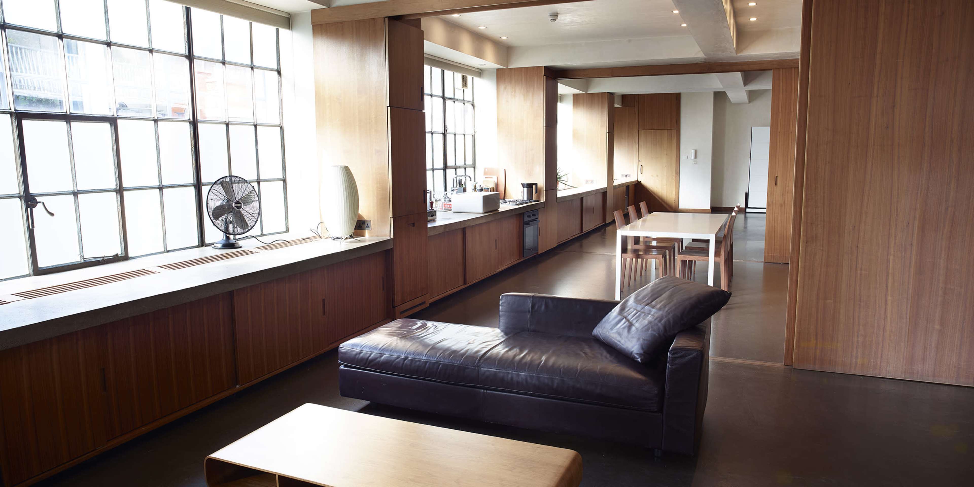 A spacious, modern interior with large windows, wooden paneling, and a seating area featuring a dark leather chaise lounge.