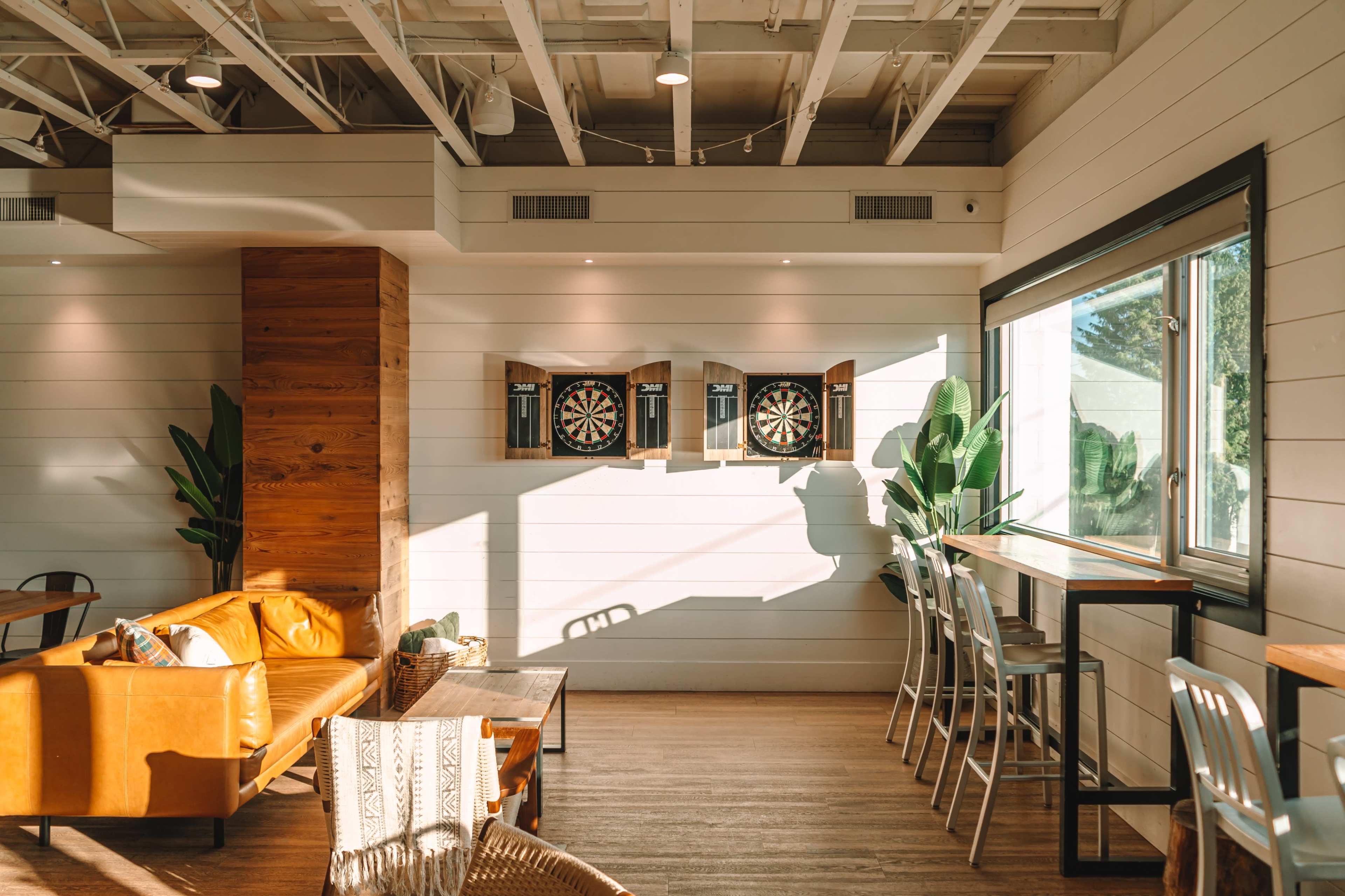 A cozy interior features a seating area with a leather couch, dart boards on the wall, and large windows allowing natural light to illuminate the space.